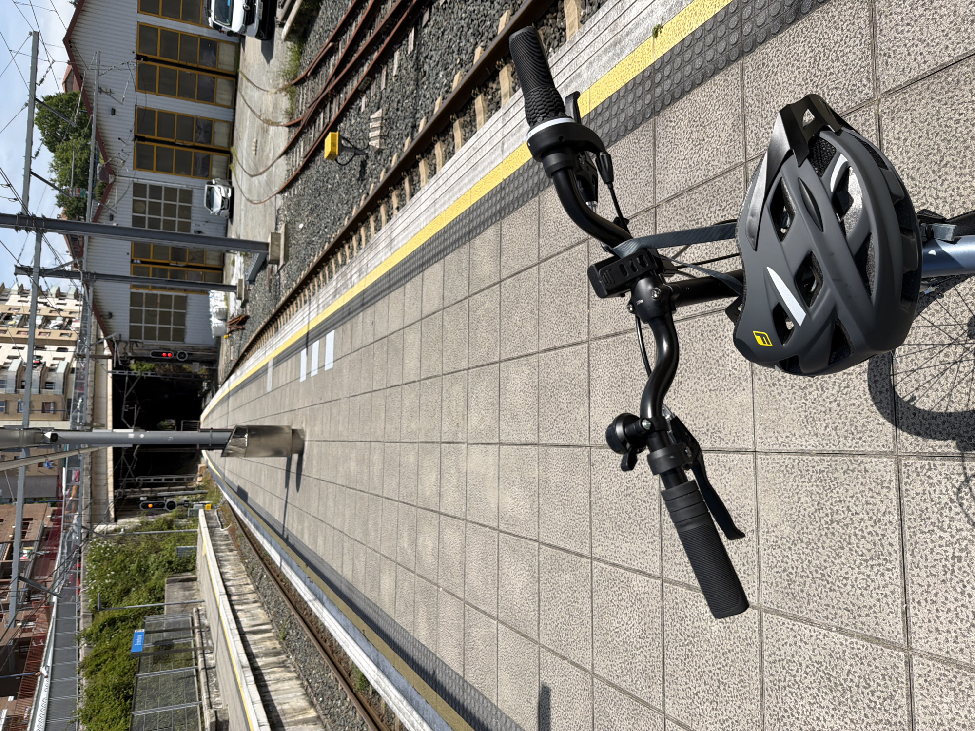 The photograph is taken from the perspective of someone standing on a train platform. The focus is on a bicycle, with the handlebars and a helmet prominently displayed in the foreground. The train platform is made of grey tiles and is lined with a yellow safety strip. To the left of the platform, there are railway tracks with ballast and sleepers. In the background, a train tunnel is visible. Adjacent to the tunnel is a building with multiple windows and a few vehicles parked outside. The surrounding environment includes other buildings and vegetation. The city of Errenteria, Spain is not explicitly identifiable from the visual information provided.