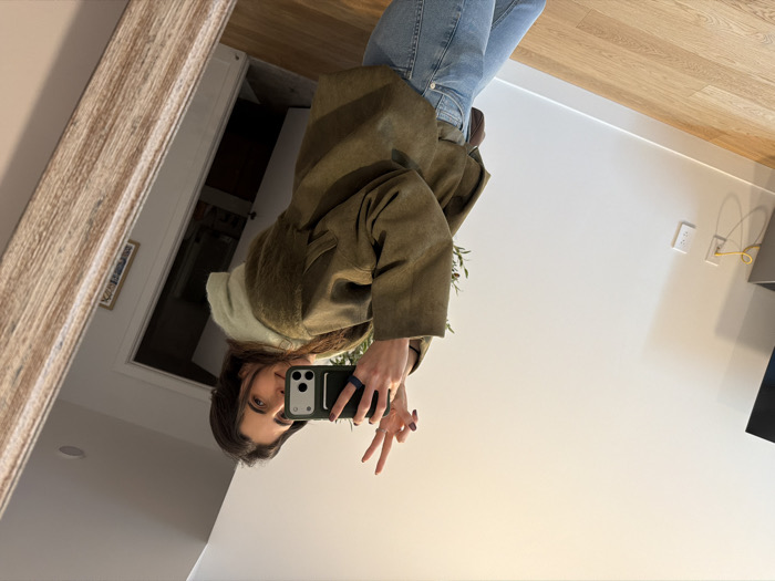 The image depicts a female individual taking a mirror selfie inside a modern residential interior. The precise location within New York City, United States of America, is not discernible from the visual content.
The subject, a female with long dark brown hair, is captured in reflection, holding a dark-cased smartphone with a visible blue ring-like grip in her left hand, pointed towards the mirror. Her right hand is raised, forming a "peace" sign with the index and middle fingers. She is dressed in a light green hooded sweatshirt beneath an olive-green or brown-grey jacket and light blue denim jeans. Her fingernails are painted with dark polish.
The environment visible in the reflection consists of light-colored walls and a light-toned hardwood floor. To the left, a dark media console supports a television screen, and a white electrical outlet on the wall has a coiled yellow cable plugged into it. Green foliage from a plant is partially visible behind the subject's left shoulder. A doorway in the background reveals a dark door handle and a framed item on the wall beyond. The mirror itself features a light-colored, possibly distressed, wooden frame on the right side of the image. A recessed light fixture is visible in the ceiling's reflection.