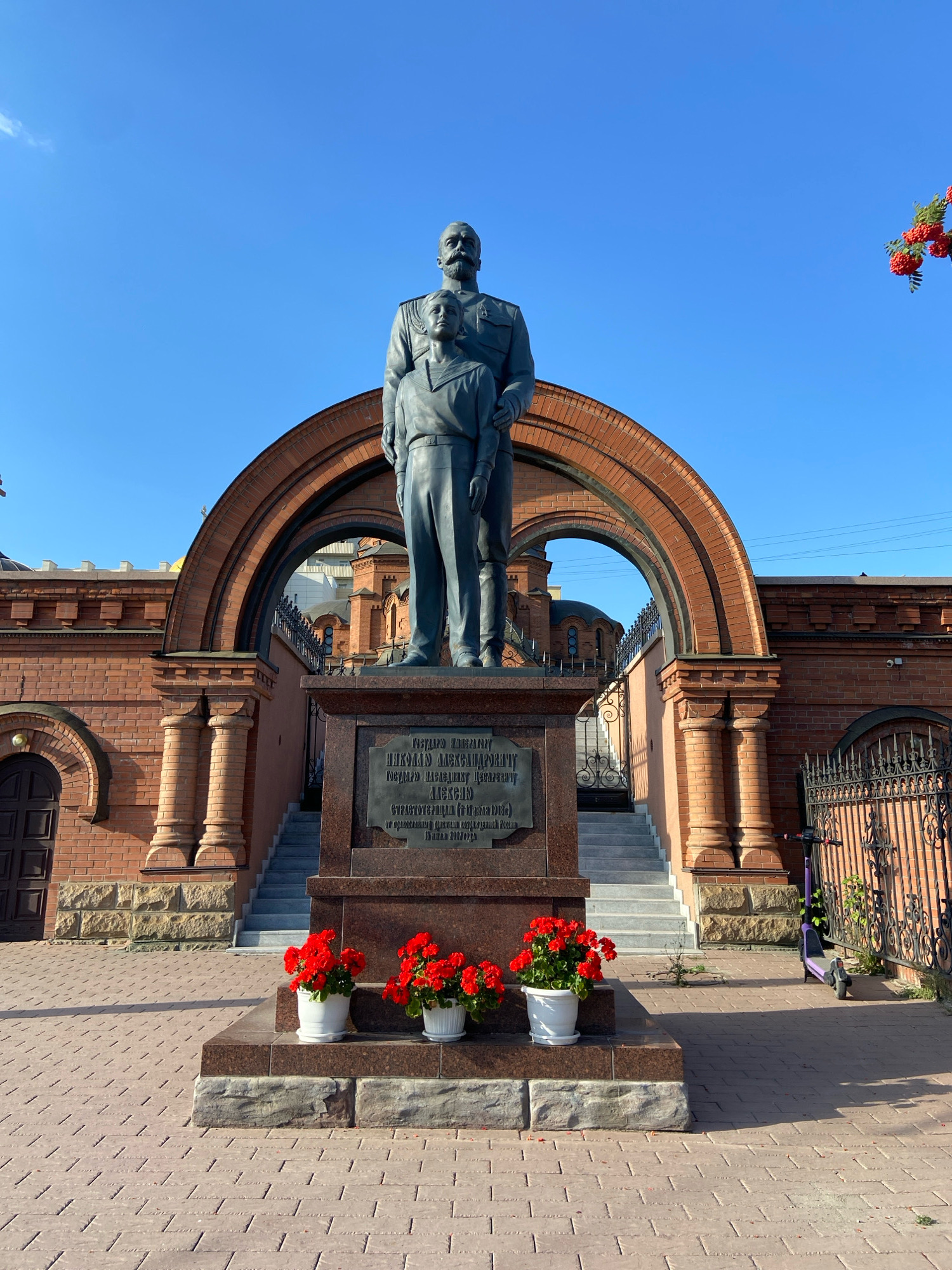 The image depicts a monument to Nicholas Alexandrovich Romanov and his son, Alexei Nikolaevich, located in Novosibirsk, Russia. The monument, cast in dark metal, features Nicholas II standing with his young son Alexei. They are positioned in front of a brick archway that leads to a staircase. The base of the monument is made of dark granite with an inscription plate. Red flowers in white pots are placed at the foot of the monument. The surrounding architecture is brick with arched windows and doorways, and a decorative metal fence is visible to the right. A purple electric scooter is parked near the fence. The sky is clear and blue, indicating a sunny day.