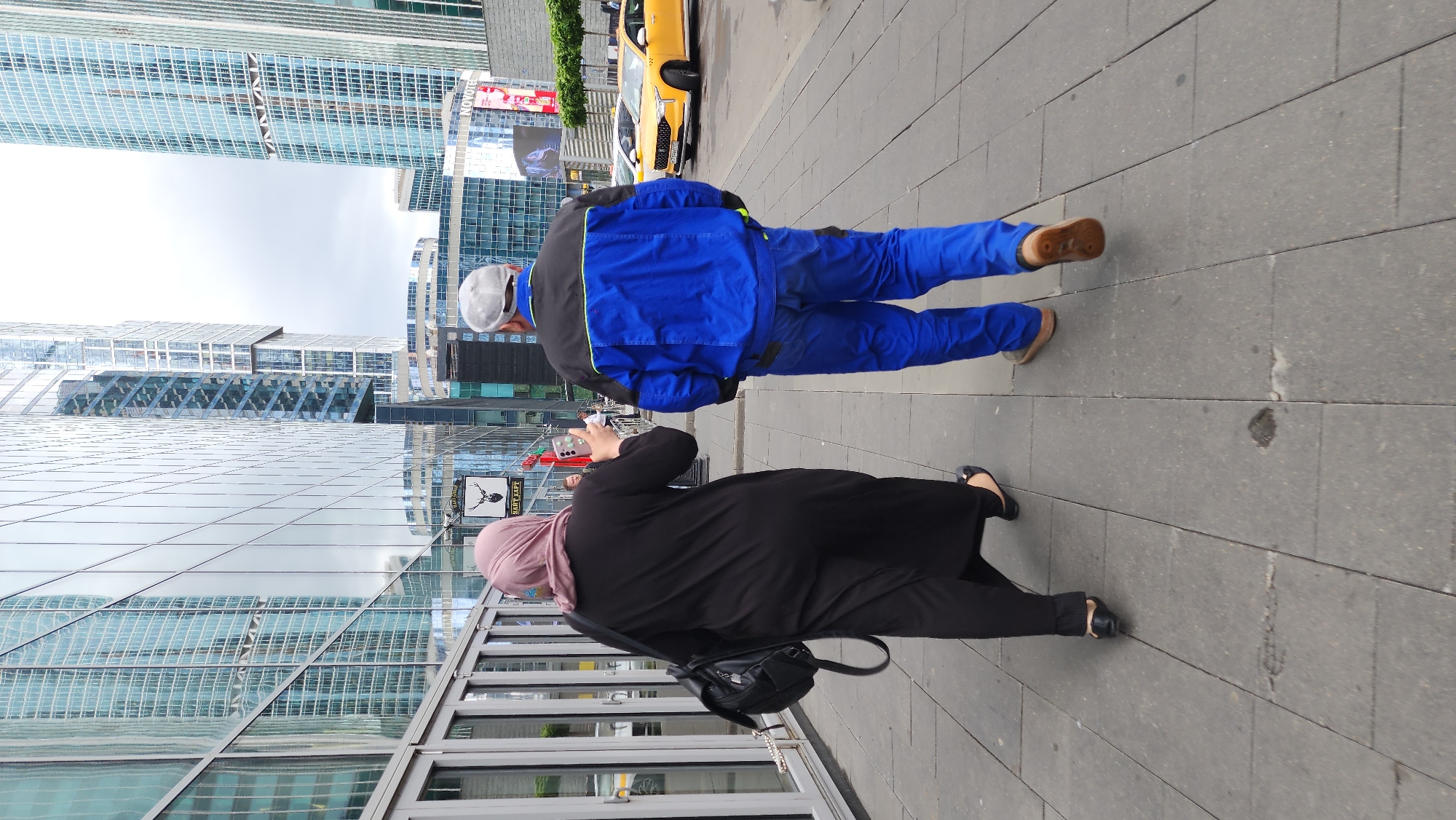 Two individuals walk on a paved sidewalk next to a building with mirrored windows reflecting nearby skyscrapers. One person, wearing a pink head covering and black clothing, holds a smartphone and appears to be taking a picture. The other person, clad in a blue work suit and cap, walks alongside. A yellow vehicle is partially visible in the background, and the setting appears to be in Moscow, Russia, judging by the architecture.