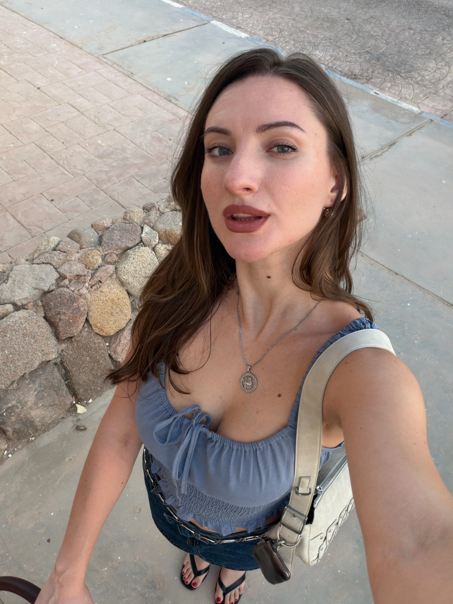 A woman is taking a selfie outdoors in Sharm El Sheikh, Egypt. She is wearing a blue crop top with a tie-front detail, dark jeans, and black flip-flops. She has a silver necklace with a pendant. A beige purse with studded details is slung over her shoulder. She is standing on a paved walkway next to a wall made of large rocks. The background shows more paved walkway and hints of a blue stripe at the edge of the paving. The lighting suggests it is daytime.