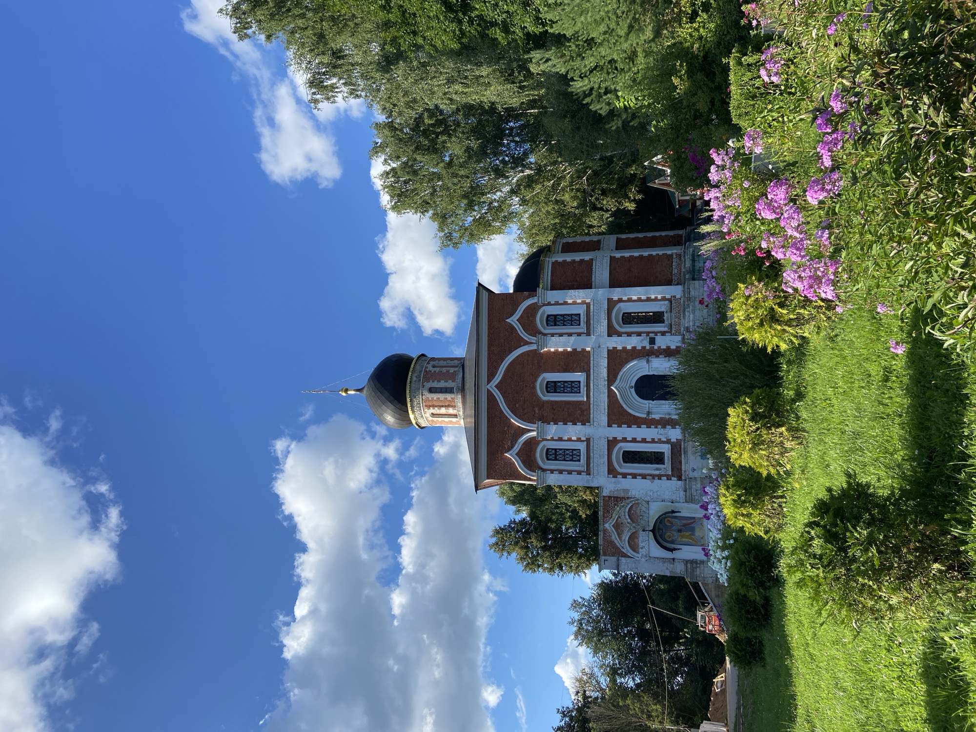 The image depicts a red brick church with a dark dome, set against a bright blue sky with scattered white clouds. The church features white architectural trim and arched windows. In front of the church, there is a garden with lush green grass and a variety of flowering plants in shades of pink and white. Trees flank the right side of the church, with one taller tree prominently visible. The ground in front of the church appears to be paved or stone. The overall scene is one of a tranquil, possibly rural, setting with a well-maintained religious building and surrounding landscape.