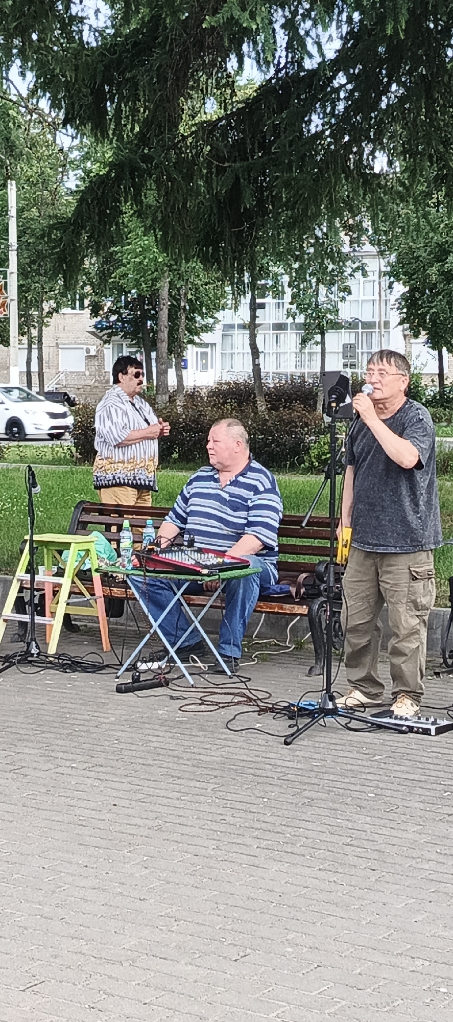 In Chaikovsky, Russia, three men are present in an outdoor setting, seemingly engaged in a musical performance. The man on the right, wearing a dark t-shirt and khaki pants, is holding a microphone to his mouth and singing. He stands near a microphone stand and some audio equipment. To his left, a man in a blue and white striped polo shirt and blue jeans is seated on a park bench. He is positioned in front of a small table holding a keyboard or mixing board and several bottles of water. A third man, dressed in a striped shirt and patterned shorts, stands to the left of the seated man and appears to be speaking or gesturing. The background consists of trees, a building with a glass facade, and a road with a white car visible. The ground is paved with interlocking bricks. The scene suggests a public performance or rehearsal in a park or town square.