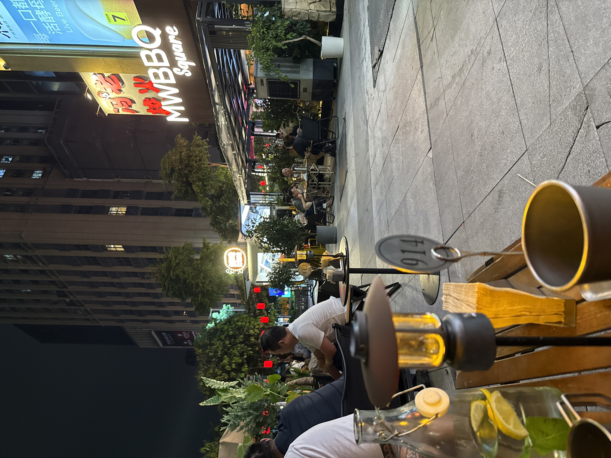 The image depicts an outdoor dining area at night, likely a restaurant or bar, in Haizhu District, China. In the foreground, a wooden table holds a glass bottle with sliced lemons and mint, a metal camping mug, and a small black lamp with a yellow light. A metal tag with the number "914" is also visible on a stand. In the midground, several people are seated at tables, some appearing to be dining and socializing. A large building with many illuminated windows is visible in the background. A prominent sign reads "MWBBQ Square" with stylized red Chinese characters above it. The overall atmosphere is of a lively evening dining establishment.