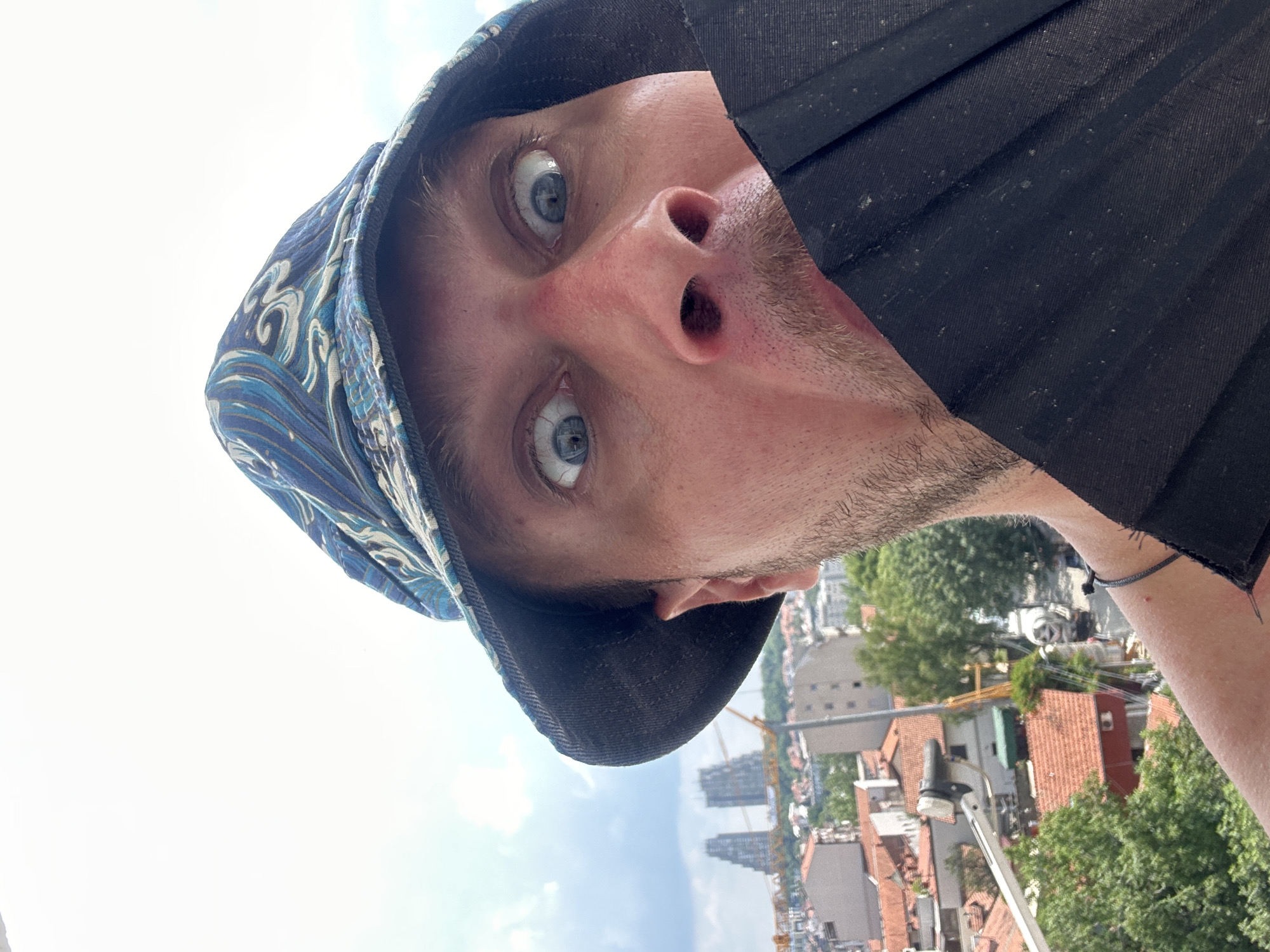 The media file is an upside-down, close-up selfie of a person's face. The subject is wearing a blue and white patterned bucket hat and a black pleated cloth over the lower part of their face, obscuring their mouth and chin. The person's eyes are wide open and looking directly at the camera, appearing startled or surprised. The background shows a cityscape with buildings, trees, and construction cranes, suggesting an urban environment. The sky is visible, indicating it is daytime. The image appears to be taken outdoors, possibly from a height, given the panoramic view of the city. The overall mood is whimsical and slightly disorienting due to the inverted perspective. The location context is Zvezdara Urban Municipality, Serbia. No visible text is present in the image.