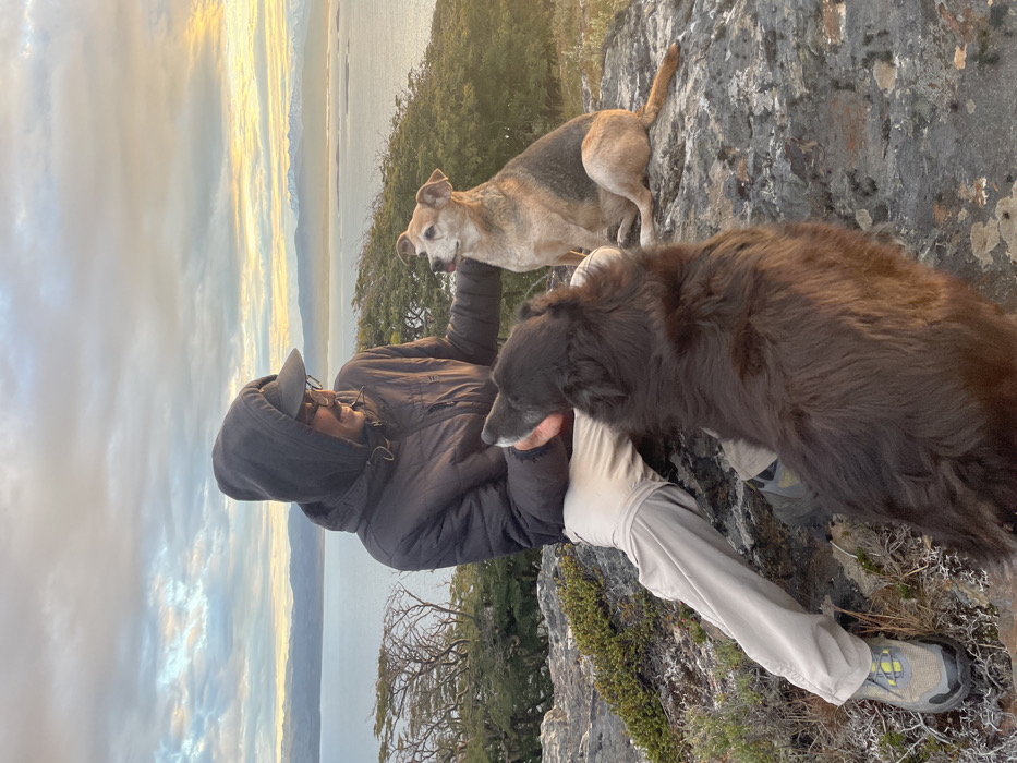A person in a dark jacket, light pants, and a baseball cap is seated outdoors. They are accompanied by two dogs; one is a tan and gray mix, standing on a rock ledge, and the other is a fluffy brown dog standing near the person. A body of water is visible in the background, with the sky displaying a mix of clouds and a golden hue, suggesting either sunrise or sunset. The landscape appears to be rocky and mountainous, covered with sparse vegetation.