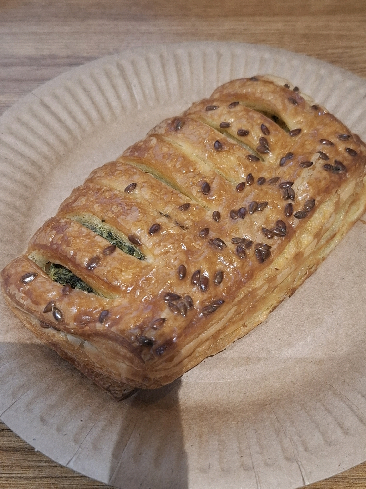 A golden-brown, flaky pastry is presented on a light brown, ridged paper plate. The pastry is rectangular with decorative cuts on top, revealing a green filling, likely spinach. Numerous small, dark seeds, possibly flax or sesame seeds, are sprinkled across the pastry's surface, adhering to a glossy glaze. The plate rests on a wooden surface, with the grain of the wood visible. The lighting is warm and overhead, casting a slight shadow. The image is a close-up shot, focusing entirely on the pastry and its immediate surroundings. There are no people or any other objects visible in the frame. The location is not specified within the image itself, and therefore, its presence in Tyumen, Russia, cannot be determined.