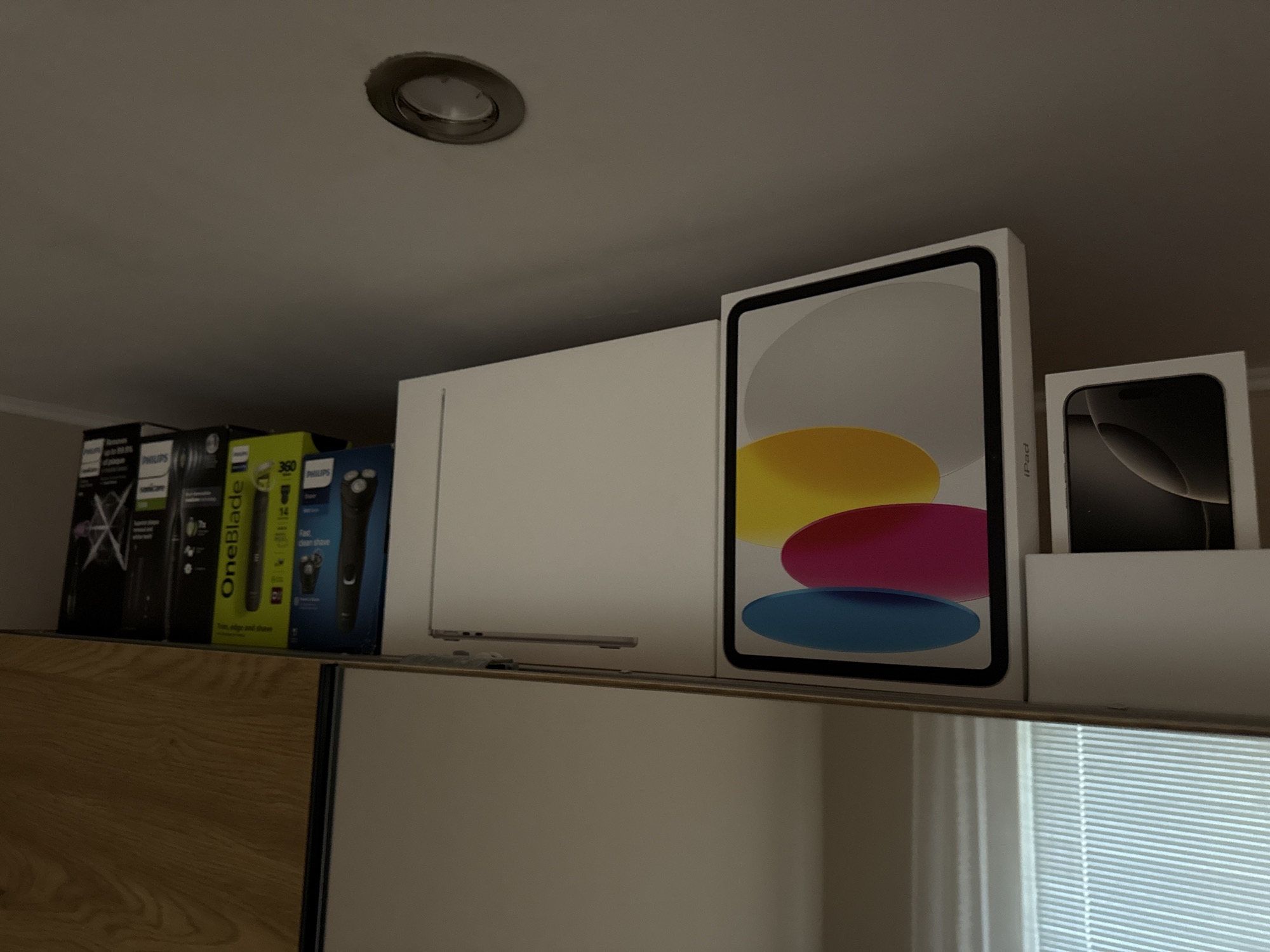 The image shows a shelf with various items. From left to right, there are several Philips OneBlade shaver boxes, a white storage cabinet, a laptop, an iPad box, and an iPhone box. The shelf is mounted in front of a window with closed white blinds. The setting is indoors. There are no people visible in the image. The location in the city of Lovech, Bulgaria, is not determinable from the image content.