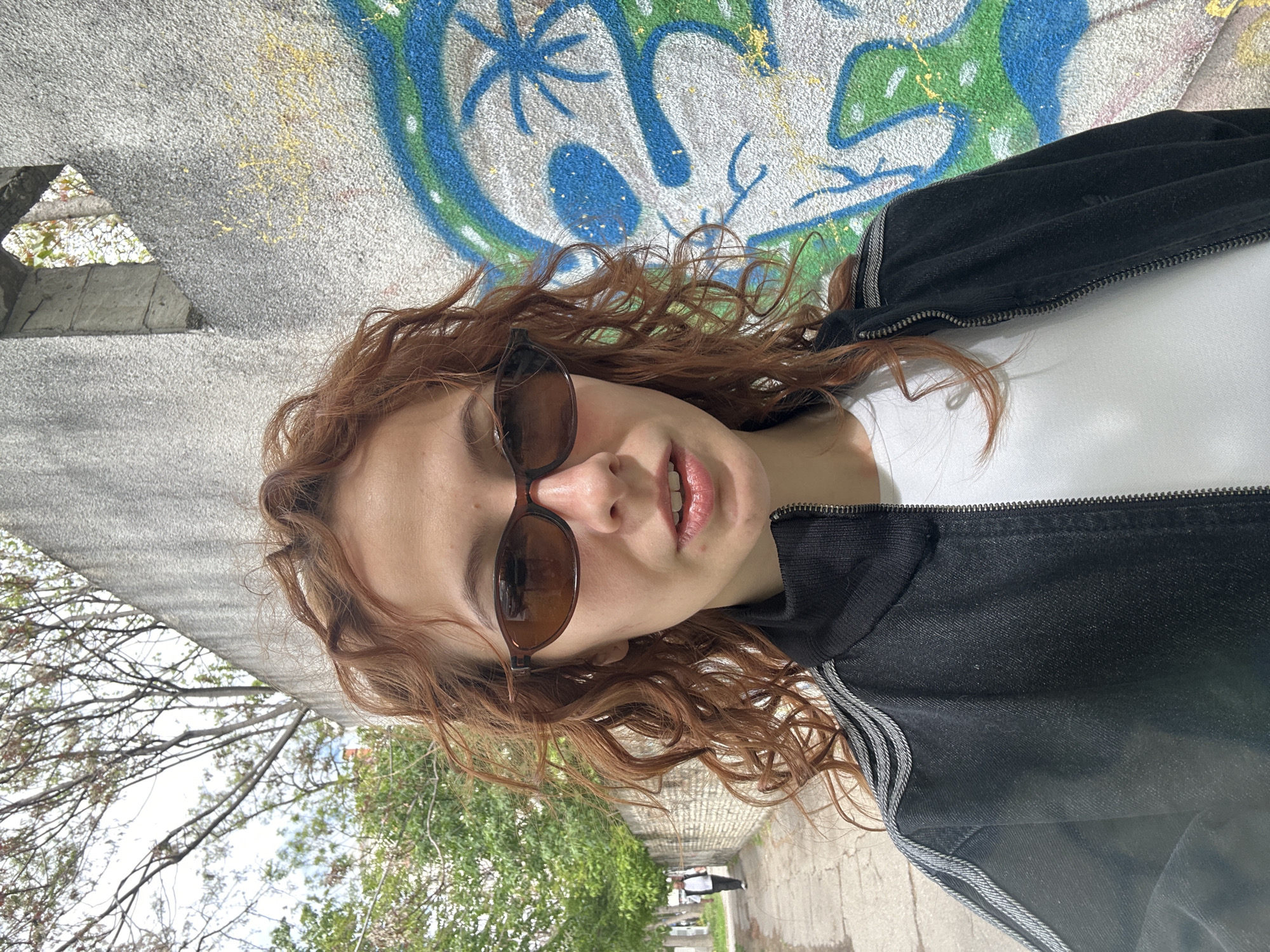 A young woman with long, wavy, reddish-brown hair is captured in a portrait-style shot, seemingly outdoors. She is wearing dark sunglasses, a white top, and a black zippered jacket featuring light-colored stripes on the sleeves. Her mouth is slightly agape, revealing her teeth.
She is positioned against a textured concrete wall, which is heavily covered in vibrant blue, green, and yellow graffiti, displaying abstract shapes, lines, and star-like patterns. To her left, partially visible through a triangular opening in the wall, are bare tree branches set against a bright sky. To her right, a paved ground surface recedes into the distance, flanked by green trees and foliage. A distant figure of a person can be seen walking in the background, further along the paved area.
The image appears to be taken during the daytime, under bright, possibly overcast, lighting conditions. The setting is an urban outdoor environment, consistent with a public space or park in Novi Sad, Serbia, as indicated by the location context. The woman's expression is neutral, suggesting a relaxed pose for the camera. No discernible text is visible within the graffiti.