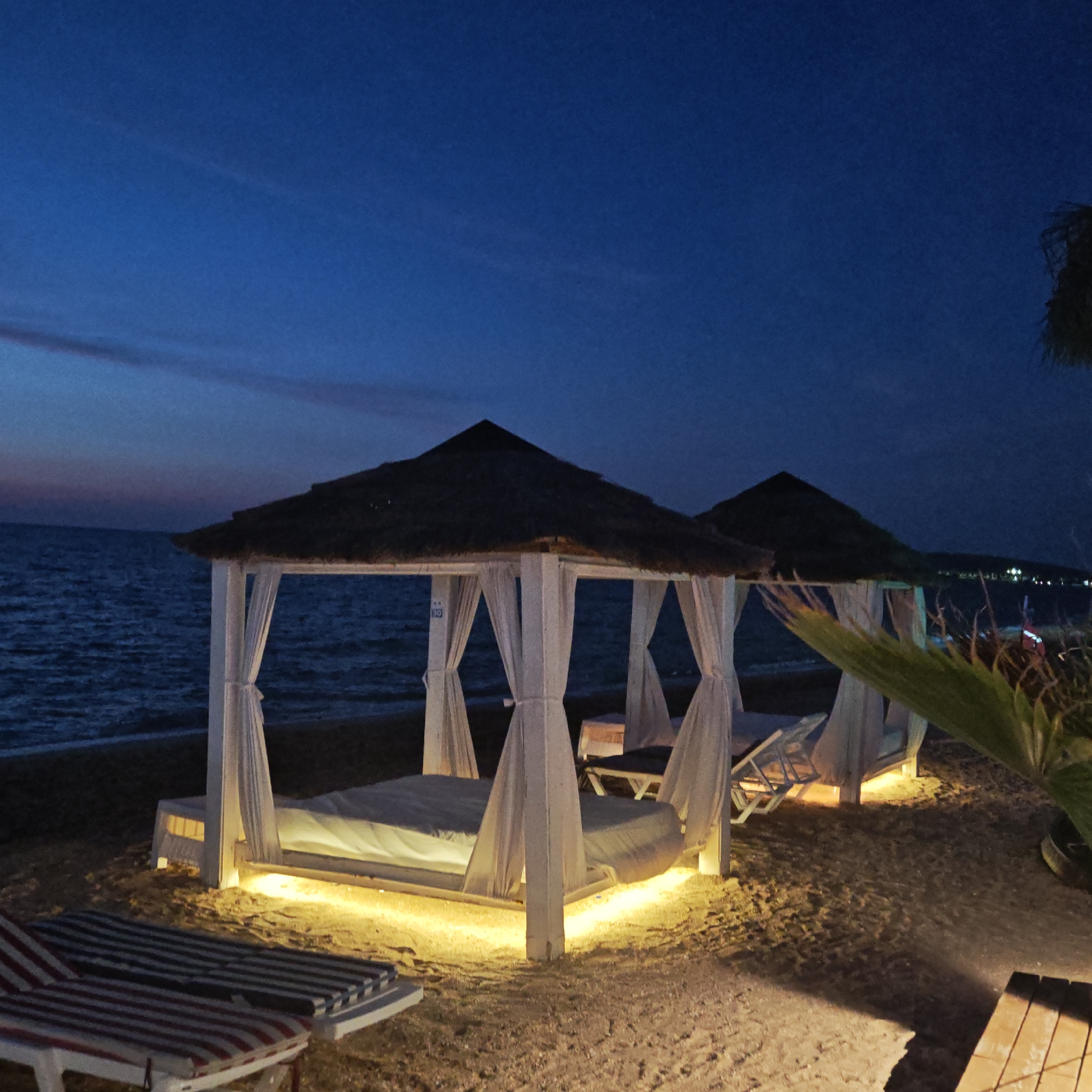The image depicts a beach scene at night in Мысовское сельское поселение, Ukraine. In the foreground, two striped lounge chairs are visible on the sandy beach. Further back, there are two open-air cabanas with thatched roofs and white curtains, each containing a bed. The cabanas are illuminated from below by yellow lights, casting a warm glow on the sand. The sea is visible in the background, with gentle waves under a dark blue sky. A palm frond is present in the right foreground, and distant lights suggest a settlement or resort area along the coast. There are no people visible in the image.