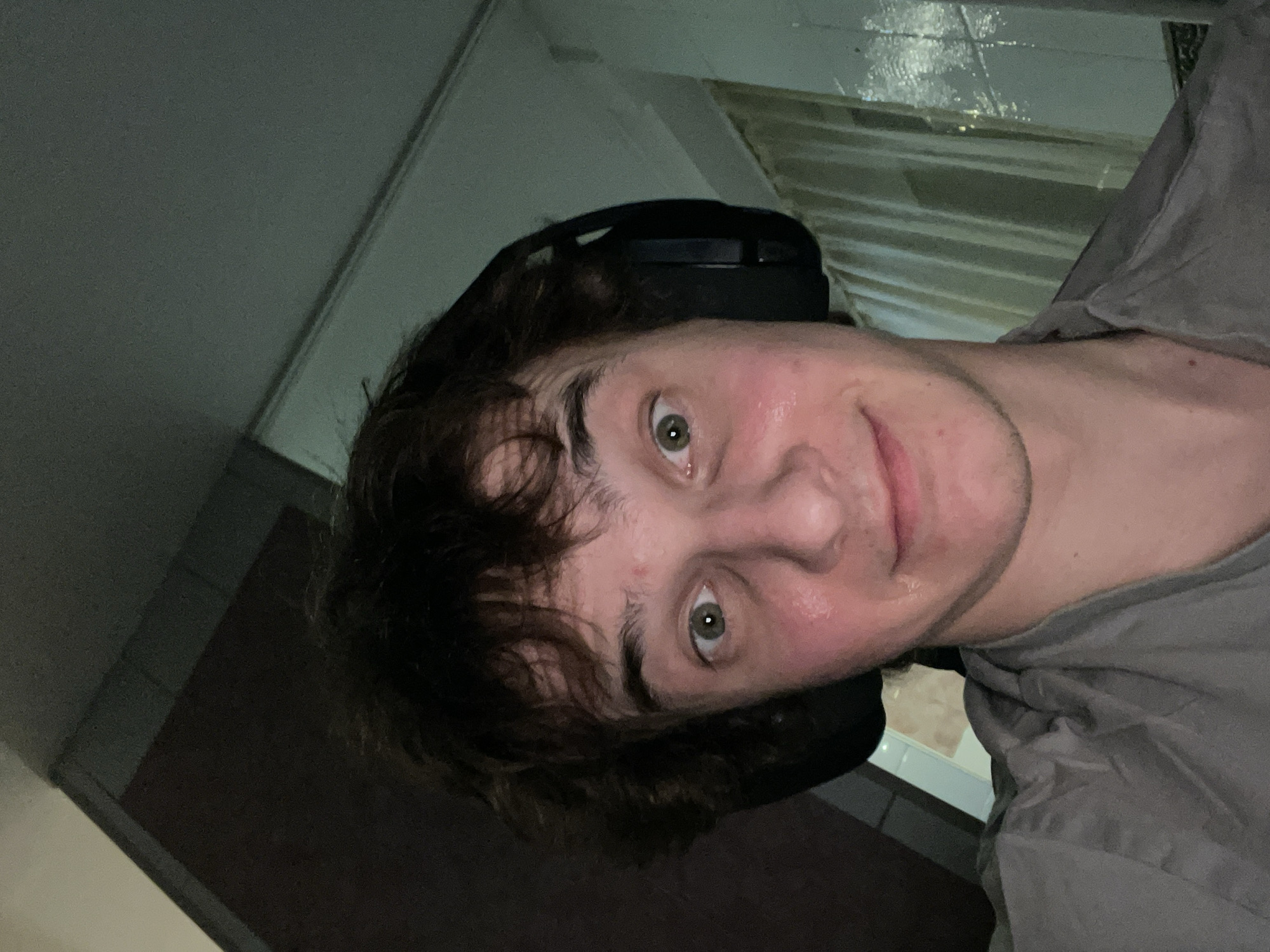 A person with brown hair and green eyes, wearing a gray shirt and black headphones, is facing the camera in what appears to be an indoor setting. The background is dimly lit, showing parts of a wall and possibly a window or reflective surface. There is no discernible evidence to place the scene specifically in Krasnoyarsk, Russia.