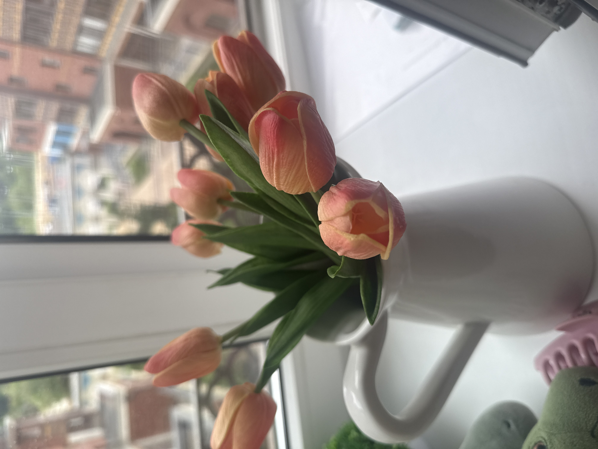 A bouquet of peach-colored tulips sits in a white ceramic pitcher on a windowsill. The pitcher has a handle on the left side. Behind the tulips, green leaves are visible. Outside the window, brick buildings with multiple windows are visible, suggesting an urban or suburban environment. The lighting suggests it is daytime.