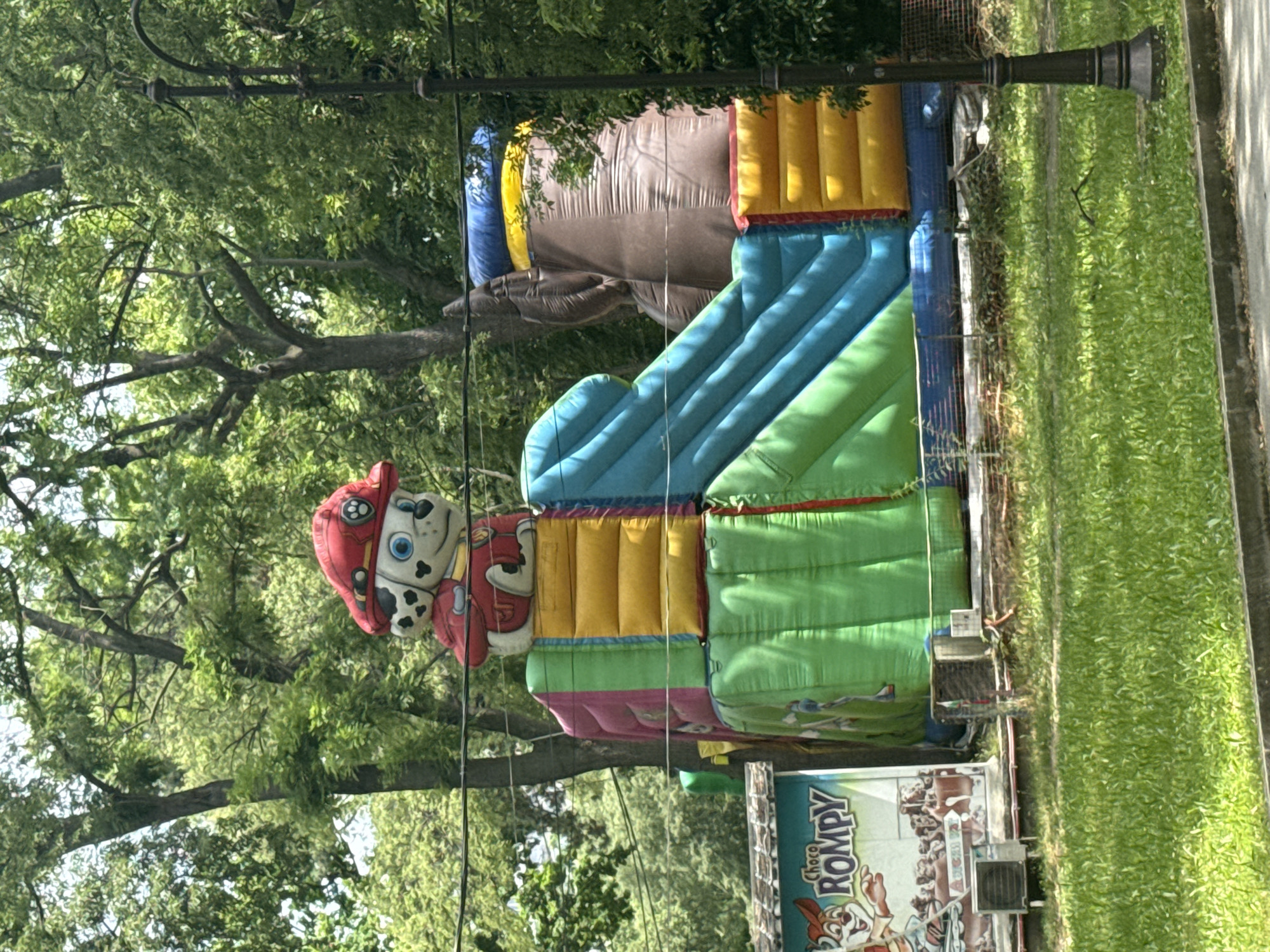 In an outdoor, possibly park setting, a large, colorful inflatable playground is set up. Atop the inflatable structure, a character resembling Marshall from Paw Patrol, wearing a red firefighter's helmet and uniform, is prominently displayed. The inflatable features multiple slides and climbing sections in vibrant colors such as blue, green, yellow, pink, and brown. To the left of the inflatable, a sign for "Choco Rompy" is visible, featuring cartoon characters and product imagery, with an air conditioning unit beneath it. The background is filled with lush green trees and foliage, suggesting a sunny day. A black lamppost stands on the right side of the image, and power lines are visible crisscrossing the upper portion. The ground in front of the inflatable is covered in green grass.