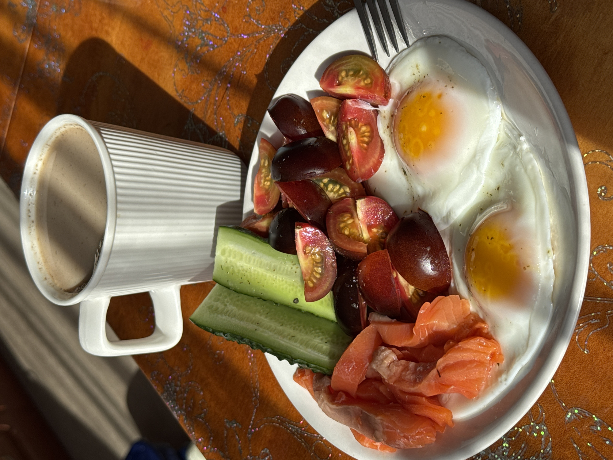 A white ceramic mug filled with a frothy, light brown beverage, likely coffee or hot chocolate, sits on a wooden surface. The mug has a ribbed texture and a modern handle. Next to the mug, on a white plate, is a breakfast spread. The plate contains two fried eggs with bright yellow yolks, a pile of thinly sliced raw salmon, sliced cherry tomatoes, and two large pieces of cucumber. A fork rests on the right side of the plate. The background is a textured orange surface with a decorative pattern of swirling lines and scattered iridescent flecks. The scene is lit by a strong light source from the right, casting distinct shadows. The overall impression is of a well-lit, possibly indoor setting, perhaps a kitchen or dining area in Stary Oskol, Russia. No people are visible in the image.