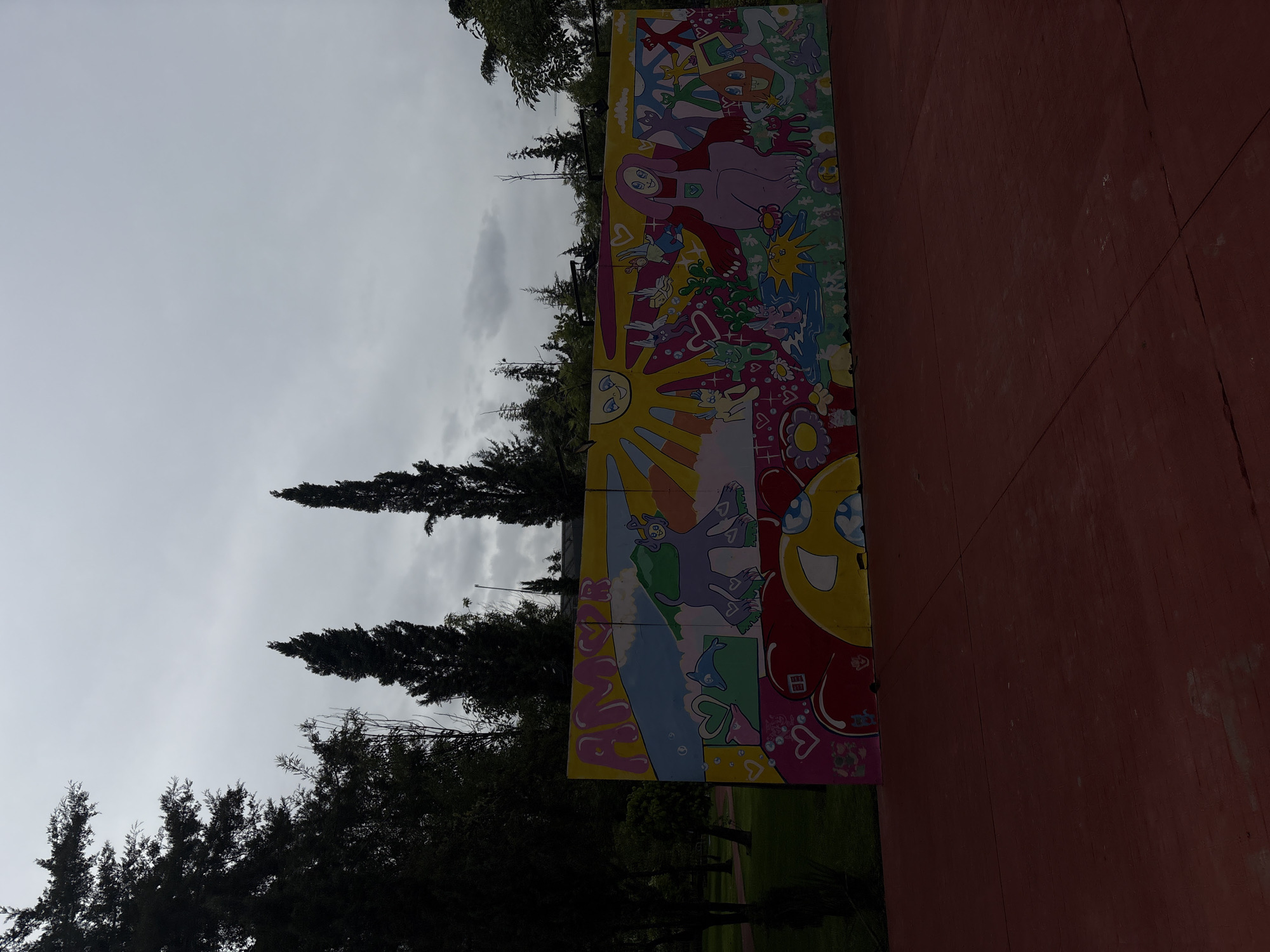 The image shows a vibrant mural painted on a long, low wall, set in an outdoor public space. The mural features cartoonish figures, hearts, flowers, and the word "AMOR" (love in Spanish). The style is whimsical and colorful, with a childlike aesthetic. Behind the mural, there are tall, dark green trees silhouetted against an overcast, gray sky. In the foreground, there is a reddish-brown paved area, possibly a park pathway or plaza. The overall impression is one of a cheerful and artistic environment, contrasting with the muted sky. While the image itself does not provide direct clues to its location, it is possible that this mural is situated in a park or public area within the city of Puebla, Mexico, known for its rich culture and art scene.