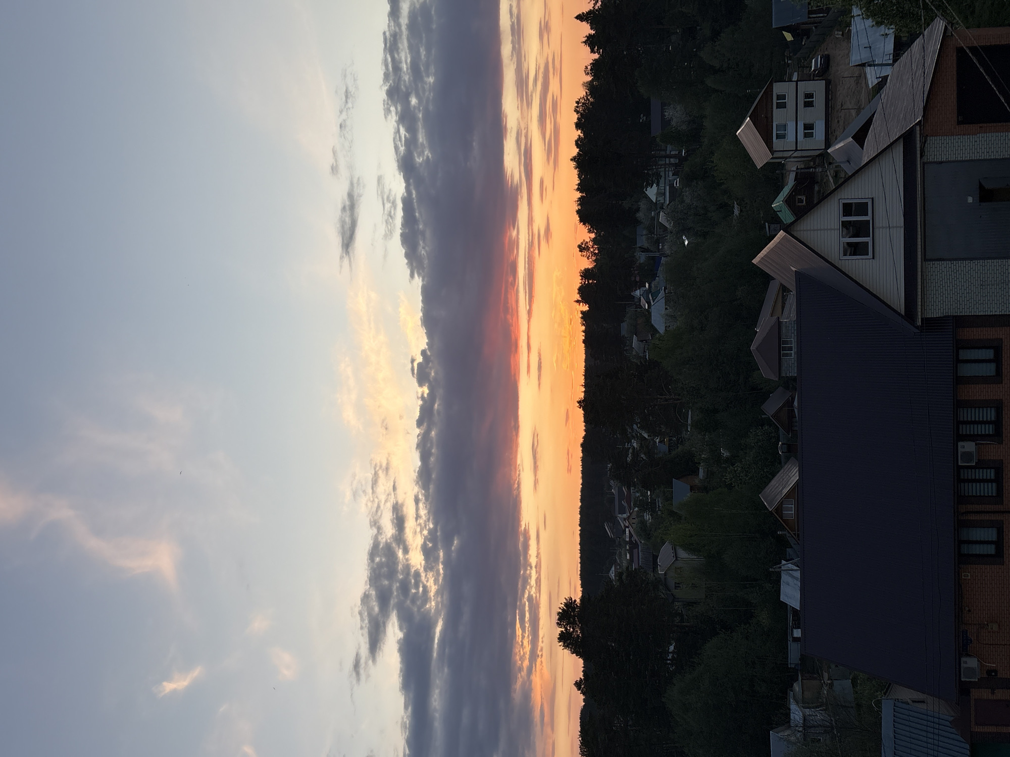 The image depicts a sunset view in Chaikovsky, Russia, overlooking residential buildings and dense tree cover. The sky is divided into thirds, the left showing light blue with thin, white clouds. The middle section displays a vertical band of sunset colors ranging from orange to purplish-grey. The right side is composed of dark, silhouetted trees and the roofs of several houses. One building, closer to the viewer, shows white siding and a dark roof with visible air conditioning units. The scene suggests a densely populated suburban or rural area near a forested zone.