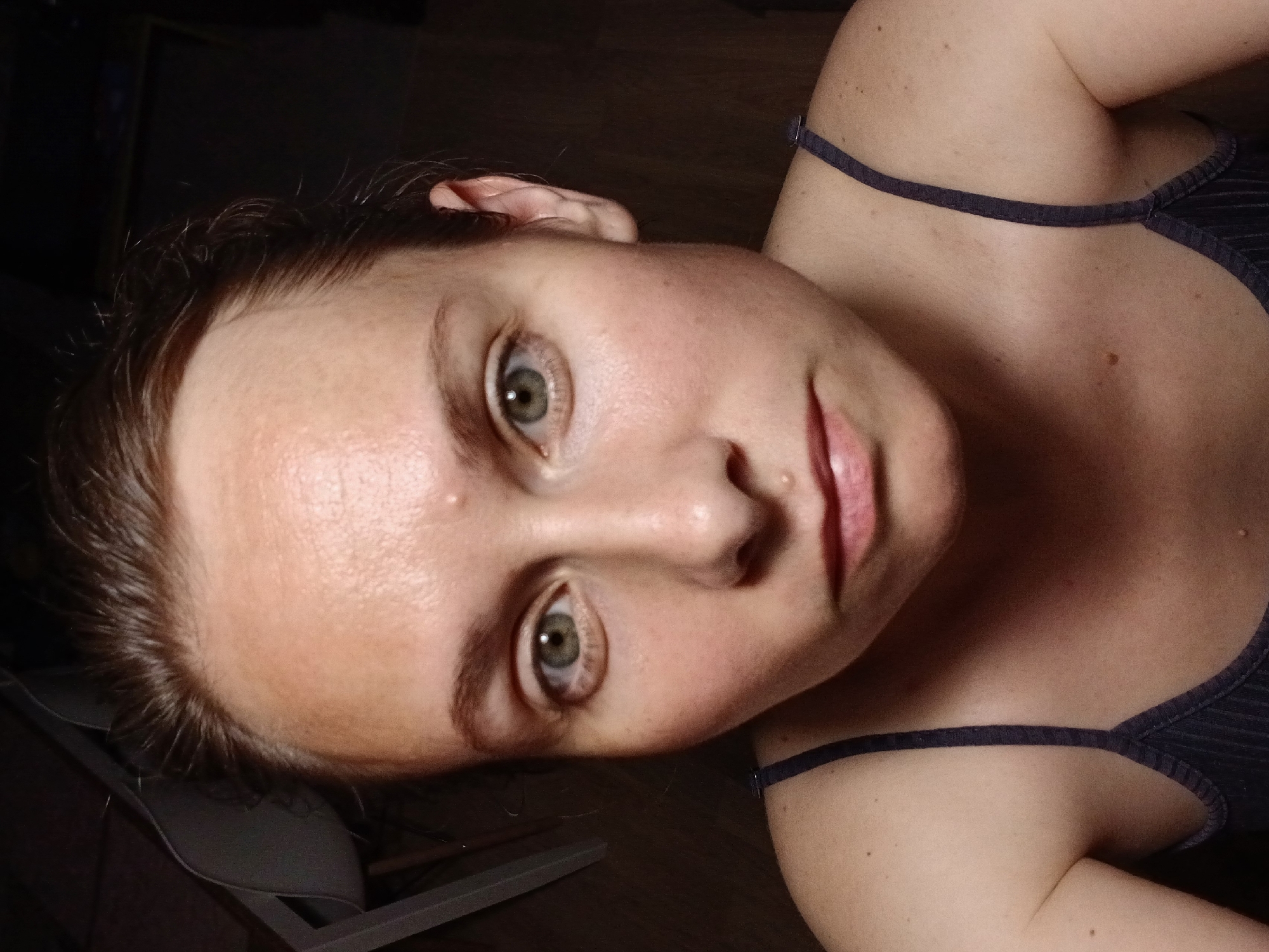 A frontal, close-up portrait of a fair-skinned woman with light brown hair pulled back, her head is tilted slightly to her left. She has green eyes, faint eyebrows, and light makeup. There is a small bump on her forehead and two small moles on her face. She is wearing a dark, ribbed tank top. The background is dark and out of focus, with a hint of a chair or furniture on the left. The lighting appears to be artificial and coming from above and slightly to the front. This image was taken indoors, possibly in a residential setting. The specific location in Yekaterinburg, Russia, cannot be determined from the visual information provided.