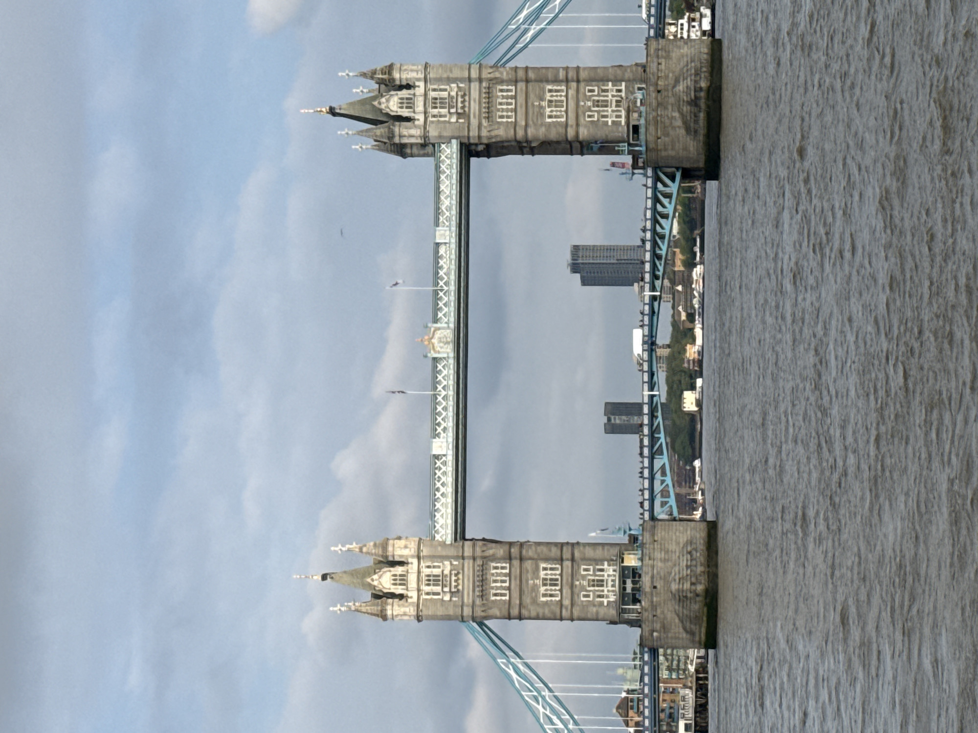 The image features a sideways view of the Tower Bridge in London, United Kingdom. The bridge's two towers are prominently visible, showcasing their Gothic architectural style with detailed stonework and turrets. The connecting walkway between the towers is also seen. Below, the River Thames appears turbulent, filling the right half of the frame. The cityscape, with modern buildings, is visible behind the bridge on the right side. The sky is overcast with grey and white clouds, setting a muted tone.