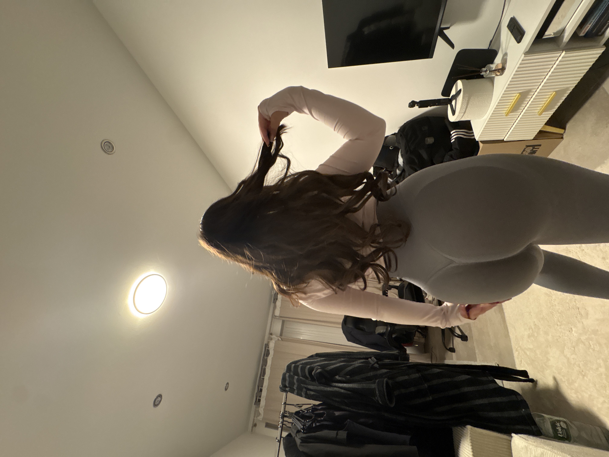 A woman with long brown hair is standing in a room, wearing gray leggings and a light pink long-sleeved shirt. She is holding a strand of her hair with her right hand. In the background, there is a television on a white cabinet with gold handles, a black backpack, a clothing rack filled with clothes, and a light on the ceiling. The location appears to be an interior space, potentially a bedroom or dressing room. No information indicates the precise location within Mönchengladbach, Germany.