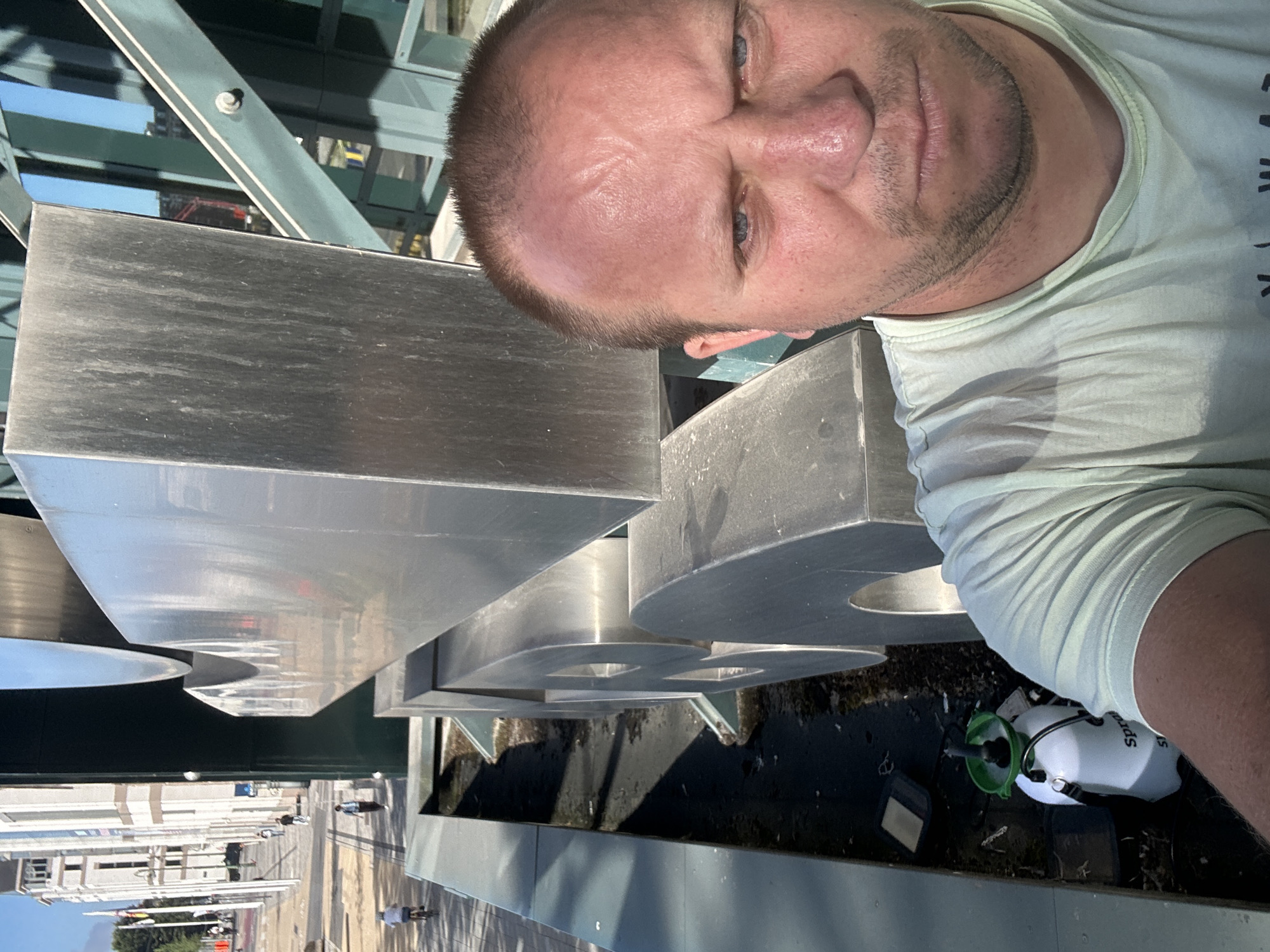 A man with fair skin, blue eyes, and short brown hair takes a selfie in front of a large, abstract, metallic sculpture in a city. He is wearing a light green t-shirt. The sculpture appears to be composed of large, curved metal shapes. In the background, to the left, is a street with a few people walking and one person on a bicycle. There are buildings and flags visible along the street. To the right of the sculpture, there is a modern-looking green and glass structure. In the lower left corner of the image, a white spray bottle with a green nozzle and handle is visible, resting on the ground next to some debris. The overall setting appears to be an outdoor public space in a city. The man's face is the most prominent subject in the foreground, with the sculpture and cityscape serving as the backdrop. The scene is lit by sunlight, casting shadows on the ground.