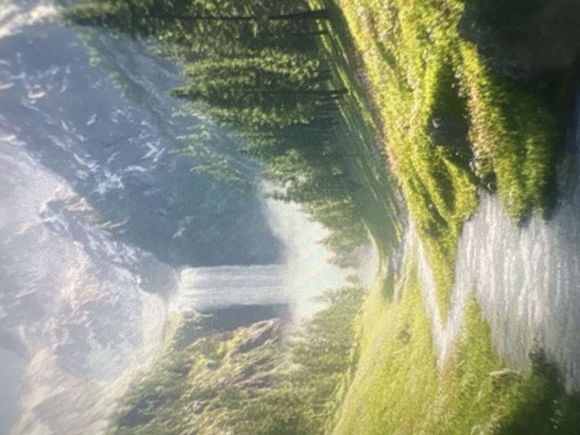 The image shows a landscape featuring a waterfall cascading down a cliff face. The cliff is covered in green vegetation, possibly moss or grass. In the background, there is a hazy view of mountains or cliffs, partially obscured by mist or fog. The composition is slightly rotated, giving a tilted perspective of the scene. The location in Kukorevo, Bulgaria is not discernible based on this image alone.