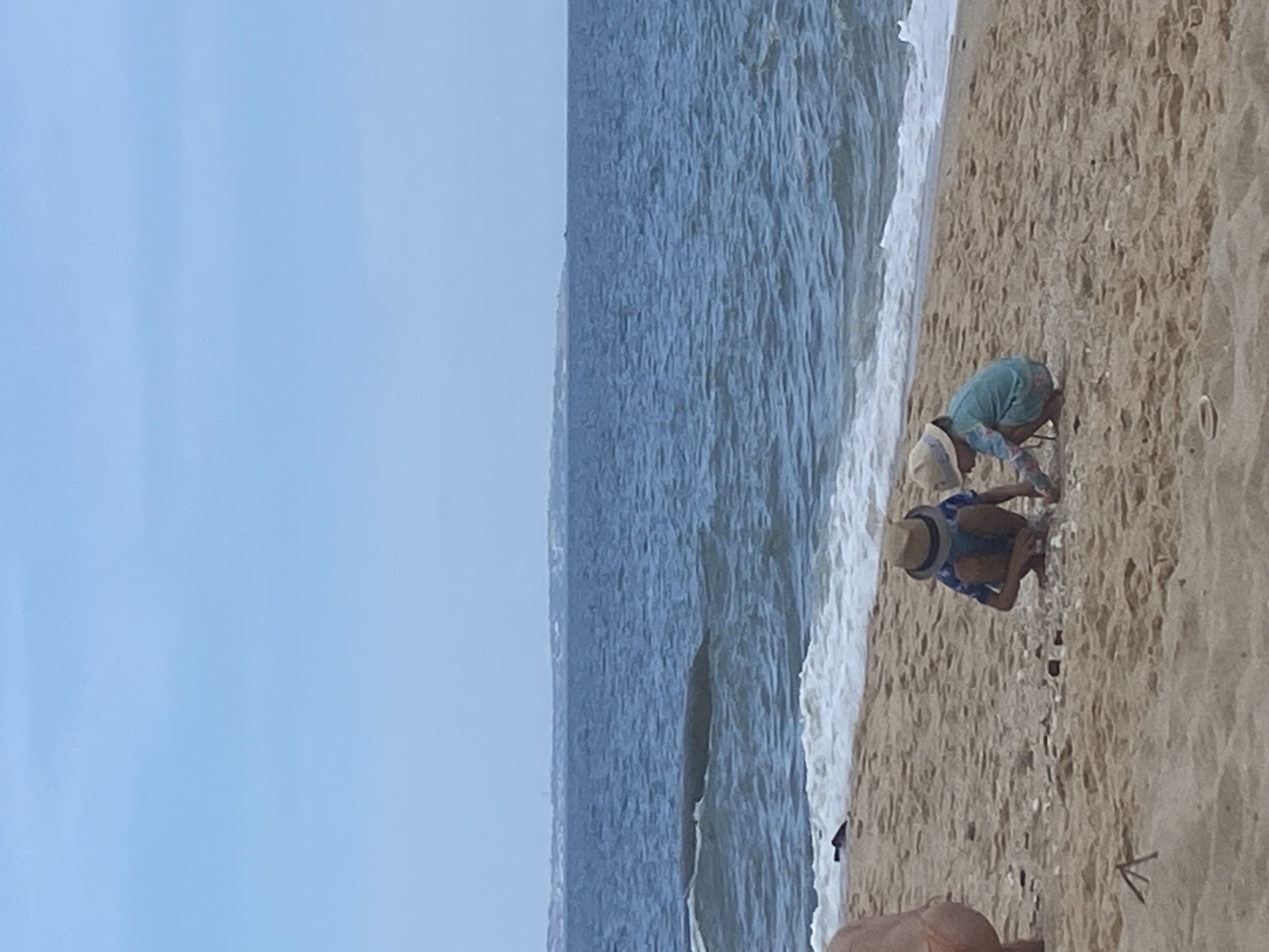 Two young children, one wearing a blue shirt and straw hat, the other a teal rash guard and straw hat, crouch on a sandy beach in Mũi Né, Vietnam. They appear to be playing with sand and shells. In the background, the ocean stretches to the horizon under a pale blue sky. A distant landmass is visible on the horizon. A portion of an adult's lower body, wearing an orange swimsuit, is visible on the far left of the image. The tide is coming in, with waves lapping at the shore.