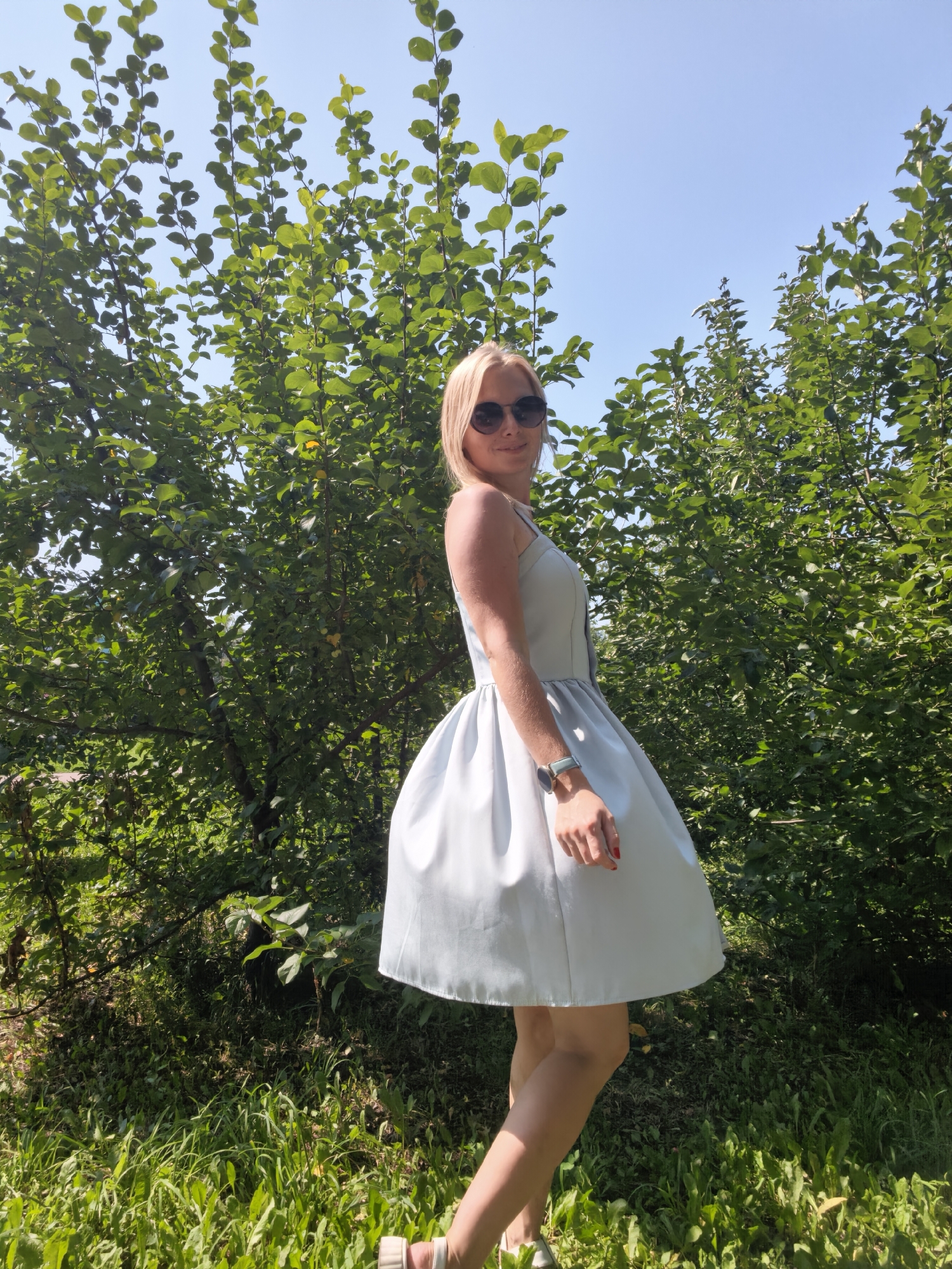A blonde woman wearing a light blue dress and sunglasses stands in front of a dense green bush. The woman is smiling and looking over her shoulder at the camera. She is wearing a watch on her left wrist and has red nail polish on her fingers. The bush is lush and full of leaves, and the sky is a clear blue. The ground in front of the bush is covered in green grass and some dry leaves. The overall impression is one of a sunny day in a natural setting. The image was taken in Sterlitamak, Russia.