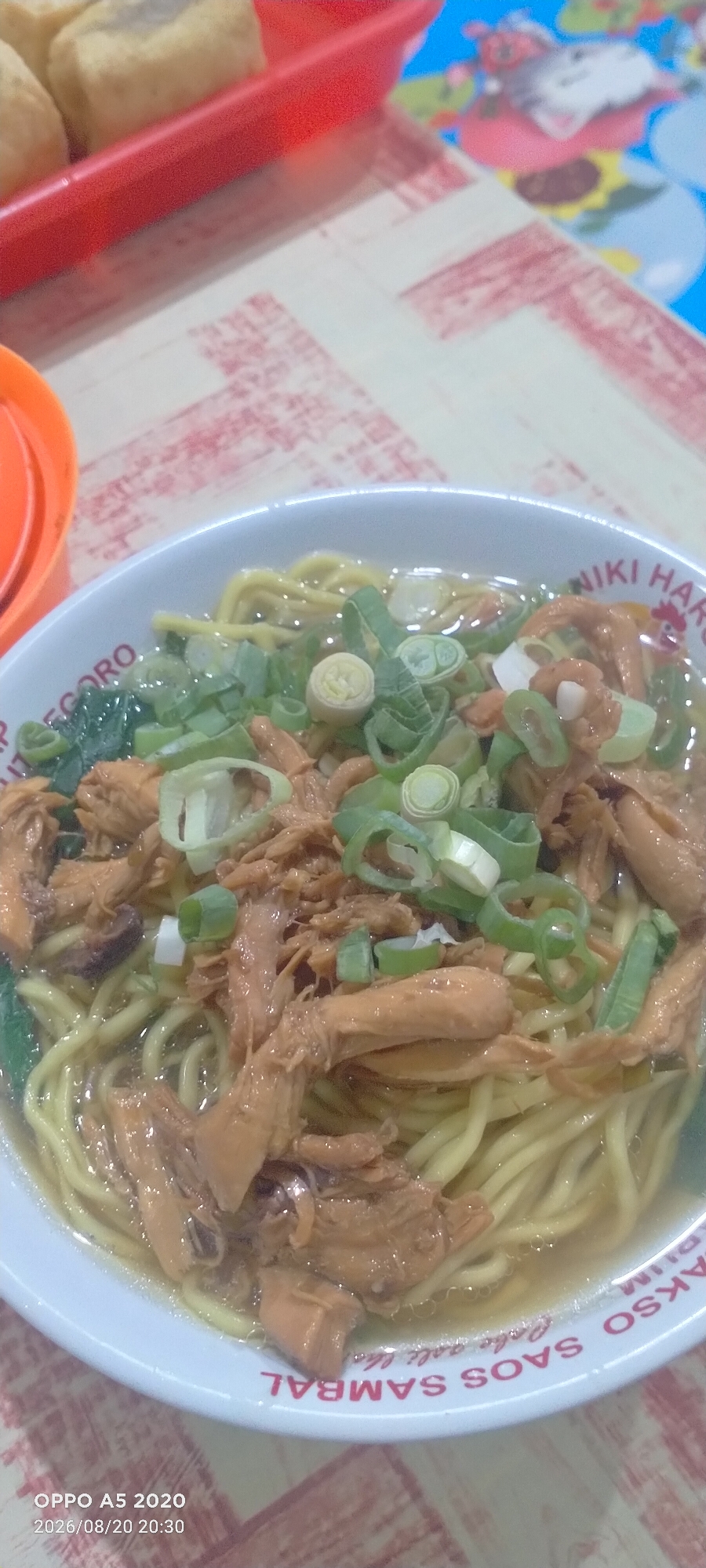 A close-up, slightly overhead shot captures a bowl of chicken noodles, a popular Indonesian dish, presented on a red and white checkered tablecloth. The noodles, a vibrant yellow, form the base of the dish, topped with shredded chicken that appears to be savory and slightly browned. Chopped green onions are generously sprinkled over the chicken, adding a fresh contrast in color and likely flavor. The bowl itself is white with red text and a graphic of a rooster, indicating a brand or restaurant. The overall impression is of a simple, delicious meal, possibly enjoyed in a casual setting. The image also shows a red plastic container in the background, and a blurry glimpse of food items inside, as well as a blue surface with colorful cartoon graphics. The timestamp "2026/08/20 20:30" and "OPPO A5 2020" are overlaid on the bottom left corner. The text on the bowl, though partially obscured, reads "WIKI HARGO", "SAOS SAMBAL", and "ASLI LE". This suggests the photo was taken in Kedung, Indonesia, a region known for its culinary offerings.