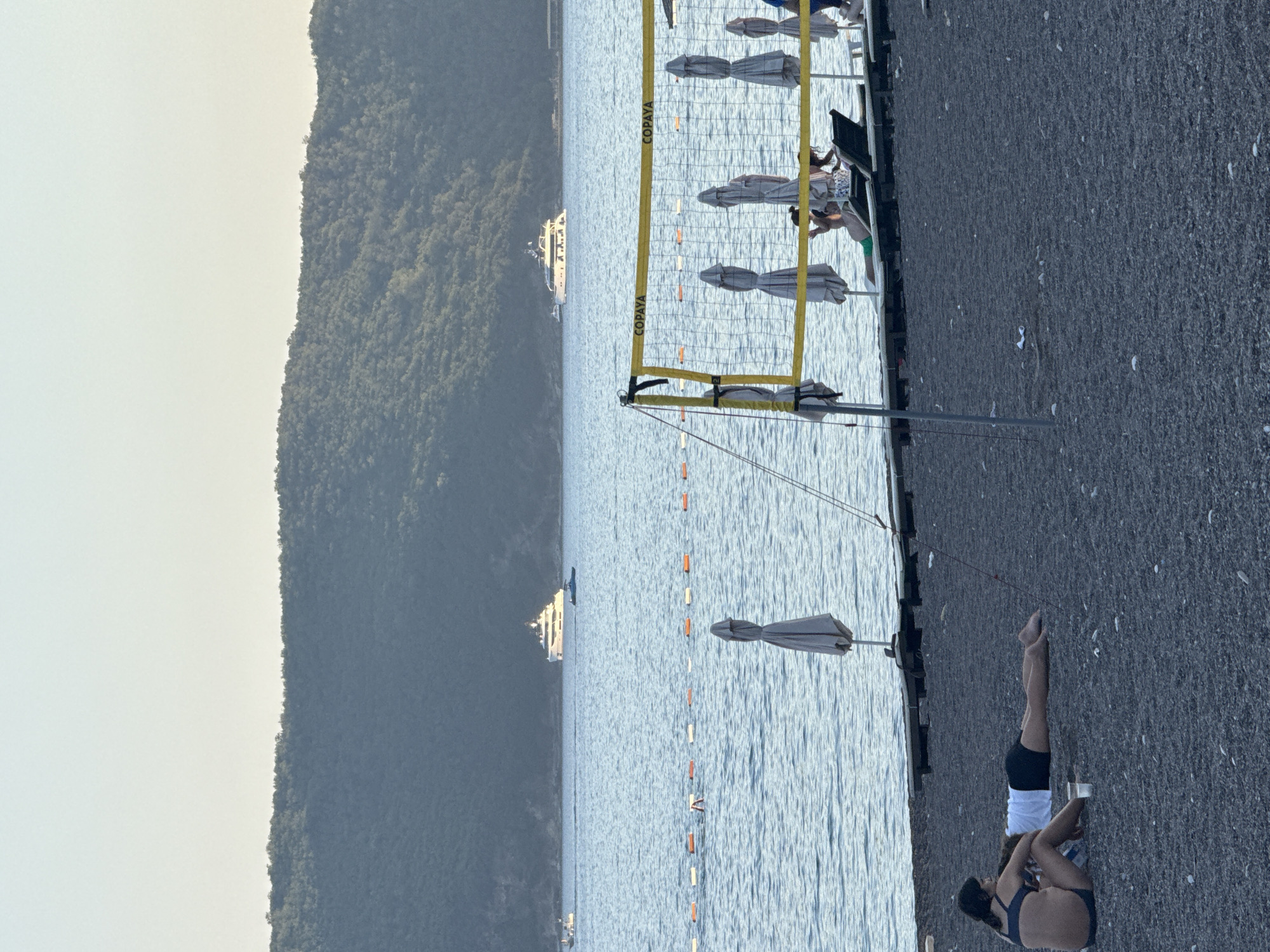 The image depicts a scenic view of the Adriatic Sea coastline in Boreti, Montenegro. In the foreground, a dark pebble beach is occupied by a few individuals. Two people are seated in the lower left corner, with one person appearing to lie down on the beach. Nearby, a clear plastic cup sits on the sand.
Further into the scene, a volleyball net with "COPAYA" branding is visible, indicating a recreational area. Behind the net, several closed beach umbrellas stand upright on a wooden deck. The sea stretches out towards the horizon, marked by a line of orange and white buoys.
In the middle ground, two large white yachts are anchored in the water. The background is dominated by a lush, green, tree-covered mountain range, characteristic of the Montenegrin landscape. The sky above is a pale, clear blue, suggesting a pleasant day. The overall atmosphere is one of a relaxed seaside resort.