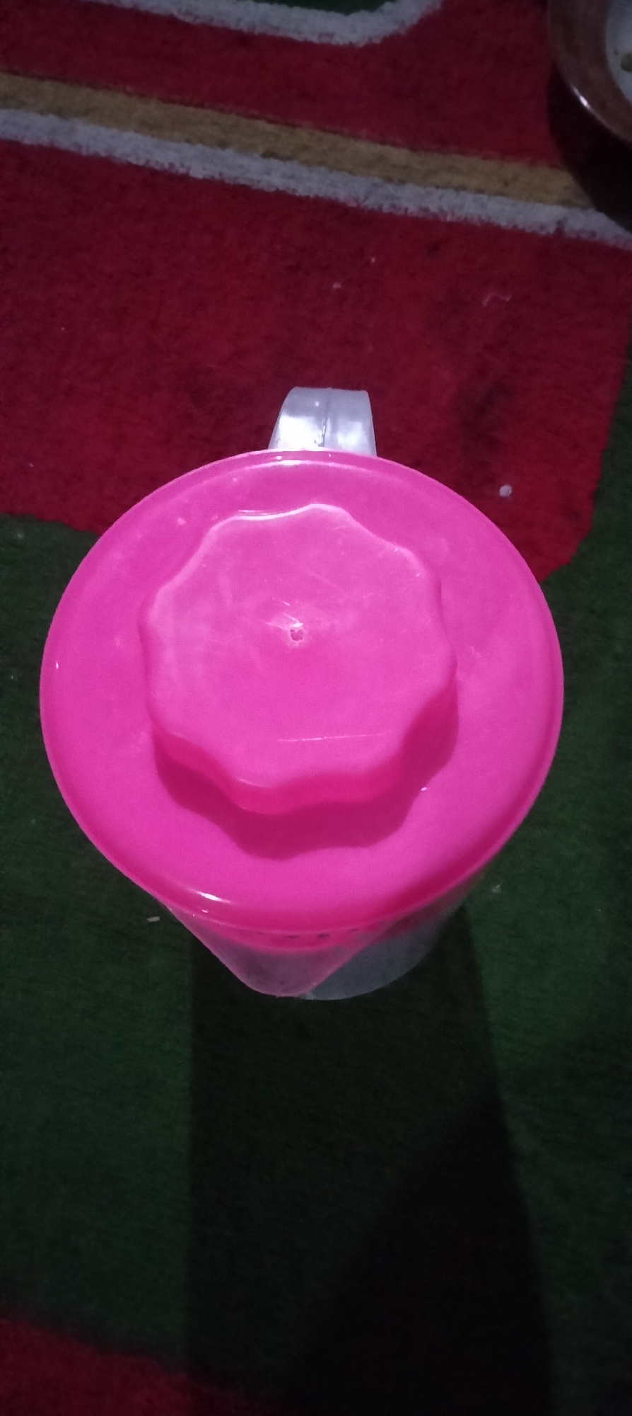 A top-down view captures a bright pink pitcher lid with a star-shaped, textured pattern in the center. A transparent, white handle protrudes from the top of the lid. The pitcher is placed on a surface that appears to be a rug with alternating stripes of red and green. The lighting suggests an indoor setting. The image shows no people or specific landmarks to identify its location in Serut, Indonesia.