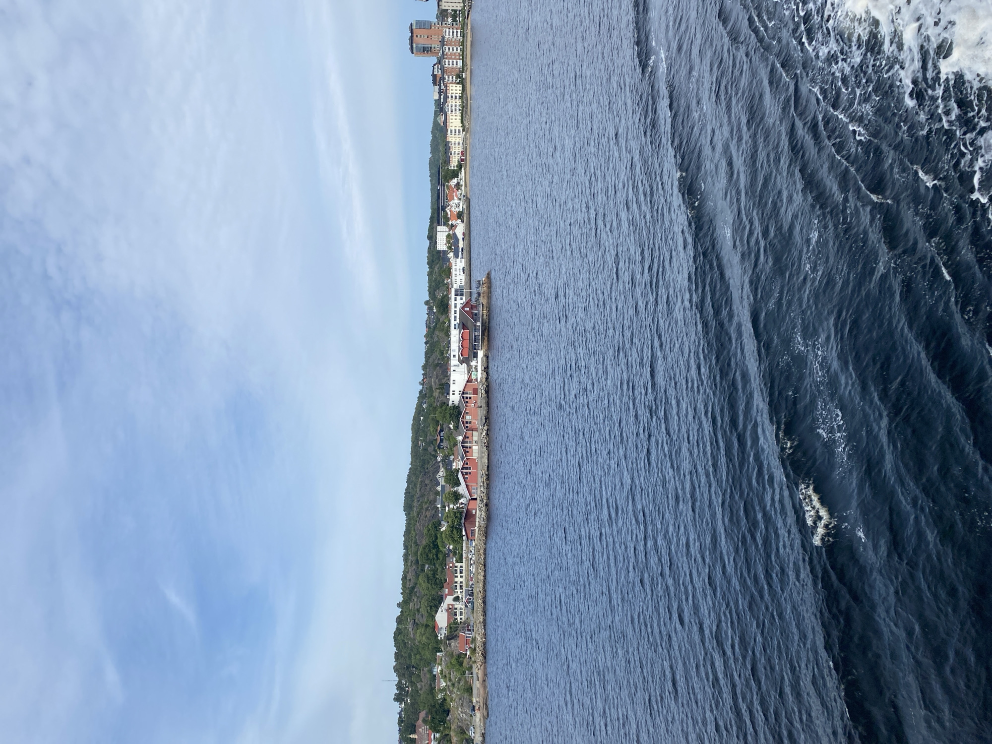 This is a wide-angle, high-angle, horizontal shot of a coastal town in Larvik, Norway. The town features a mix of residential buildings, some with red roofs and white walls, and taller, modern buildings. The town is nestled against a lush green hillside dotted with trees. A road runs along the coastline, with parked cars visible.
The scene transitions sharply from the town to a deep blue, choppy body of water. The water on the right side of the image is turbulent, with white foam and distinct wave patterns, suggesting movement or a wake. The sky above is a light blue with wispy clouds, indicating fair weather. The time of day appears to be daytime, likely late morning or early afternoon, given the brightness and the angle of the light.
There is no visible text in the image. The overall impression is of a scenic coastal landscape, likely captured from a moving vessel, given the perspective and the water's motion. The emotion conveyed is one of peaceful observation of a well-populated coastal area.
