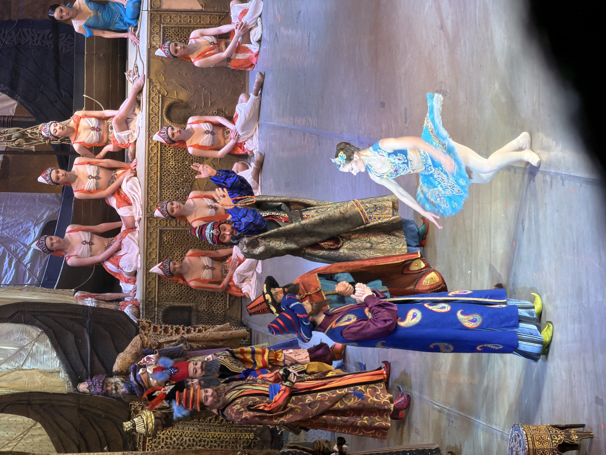 The image captures a stage performance, presumably ballet, featuring multiple performers in elaborate costumes. The scene includes dancers dressed in harem-style attire and a ballerina in a blue tutu. In the foreground are performers in ornate robes and headwear. The photograph was taken in Izhevsk, Russia.