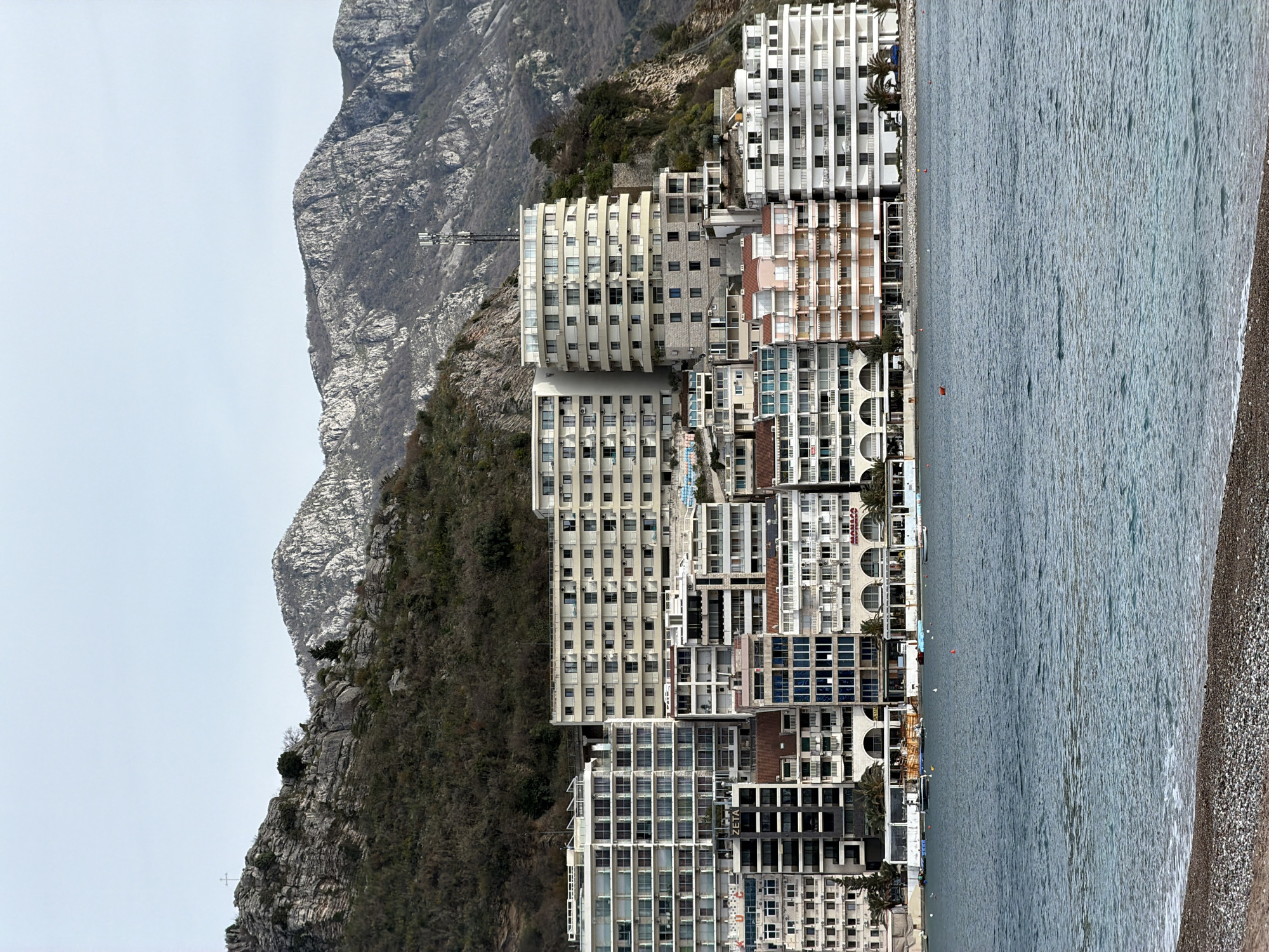 The image shows a coastal view of Rafailovići, Montenegro, with a shoreline lined by multi-story buildings transitioning towards the sea. The architecture is modern, with numerous windows and balconies. A rocky mountain formation rises in the background, contrasting with the buildings and the water. The sea appears calm with a pebbly beach. There are no people visible. The lighting is overcast.