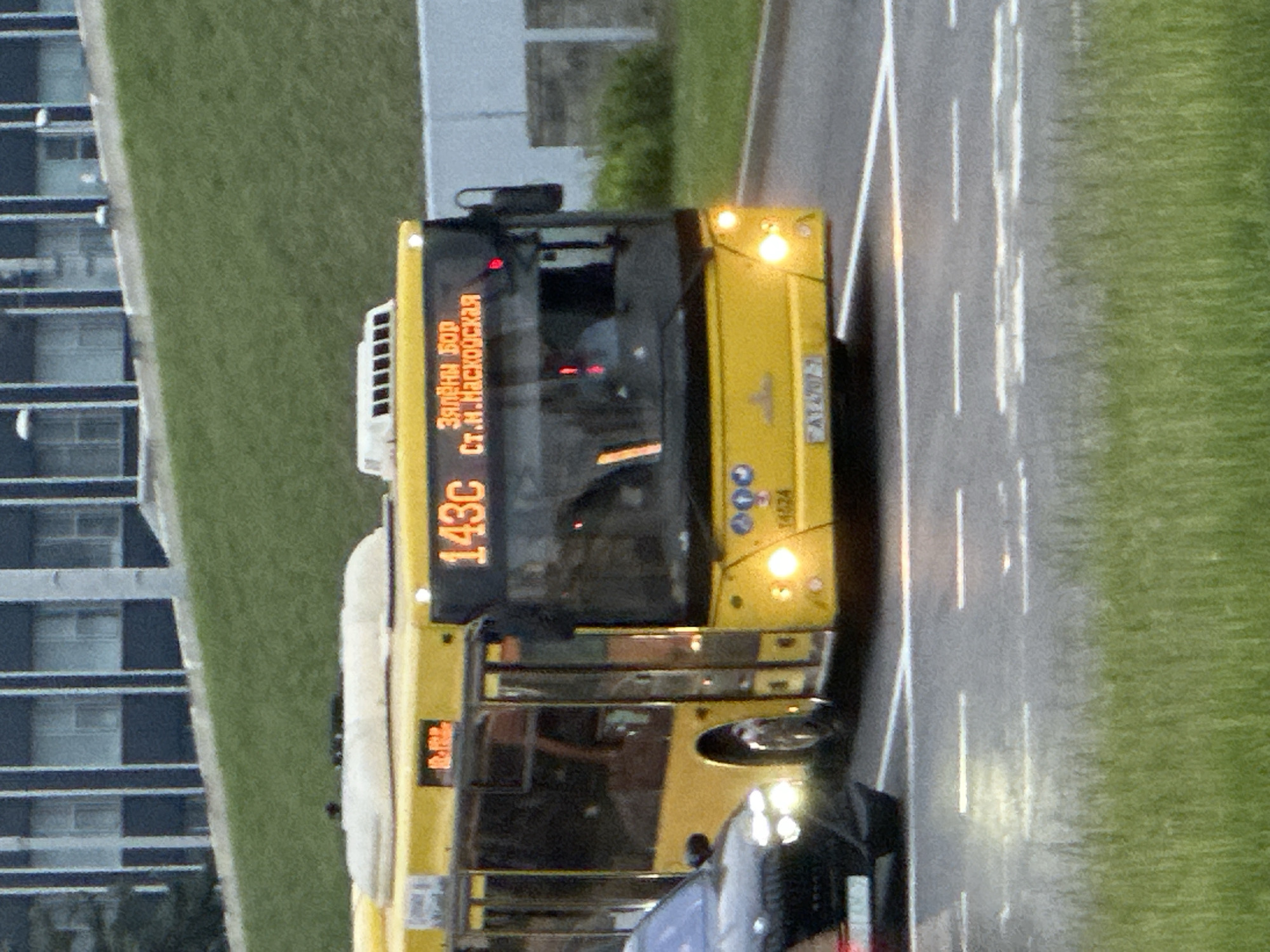The image depicts a yellow MAZ city bus, identified as route 143C, traveling on an asphalt road in Minsk, Belarus, during daylight hours, likely overcast or late afternoon. The bus's digital display indicates its destination as "ЗЯЛЁНЫ БОР" (Zialiony Bor) and "СТ.М.МАСКОЎСКАЯ" (Metro Station Maskoŭskaja). The bus model is identifiable by the MAZ logo on its front grille and an operational number "14424" on the lower right front. Its headlights are illuminated. A black BMW vehicle, possibly an SUV, is partially visible in the left foreground, its front headlights also illuminated, positioned slightly ahead and to the left of the bus.
The road features white dashed lane markings. To the immediate right of the bus, a concrete barrier or fence separates the road from a grassy area. A substantial green grassy slope occupies the mid-ground, leading up to a large, multi-story building with a dark facade and numerous windows in the distant background. No pedestrians are visible. The scene captures a typical urban transit moment in Minsk.