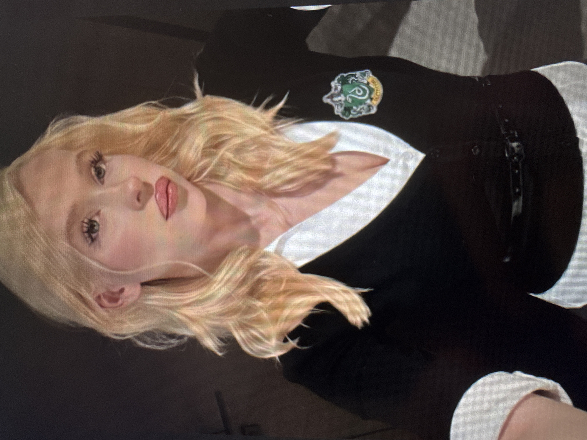 An individual, likely female, with long, wavy blonde hair and fair skin, is depicted in a portrait-style image. The image is rotated 90 degrees clockwise. She is wearing a black top or cardigan over a white collared shirt, with the white collar and cuffs visible. A distinctive green, silver, and gold emblem, strongly resembling the Slytherin house crest from the Harry Potter series, is prominently displayed on the left side of her black top. The emblem features a stylized serpent. The black garment also includes a studded strap or belt with a buckle across her mid-section.
Her face is made up with prominent eyelashes and glossy pink lipstick. She gazes directly at the camera with a neutral to slightly posed expression. The background is uniformly dark and appears to be an indoor setting without any specific details, putting the focus entirely on the subject. There is no visible text. The overall activity is posing for a photograph.