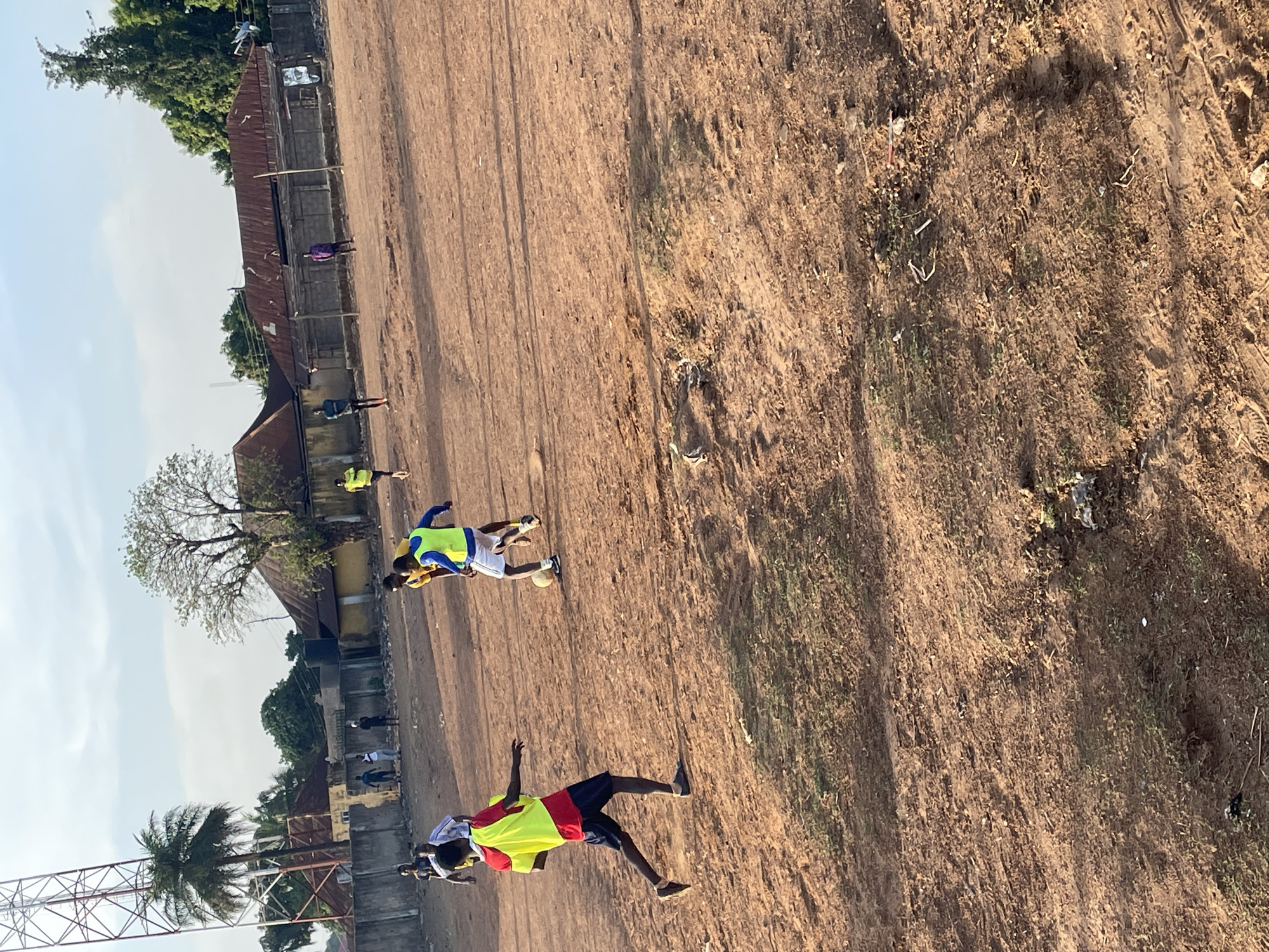 The image shows a dirt field in Akwanga, Nigeria. Two people wearing brightly colored sports vests are on the field. One person is wearing a yellow and red vest with dark shorts, and the other is wearing a yellow and blue vest with white shorts. The person in the yellow and blue vest is near a white soccer ball. Several buildings with rusted roofs and bare trees are in the background. A communication tower is also visible. The sky is a pale blue color. Additional people are visible in the background near the buildings.