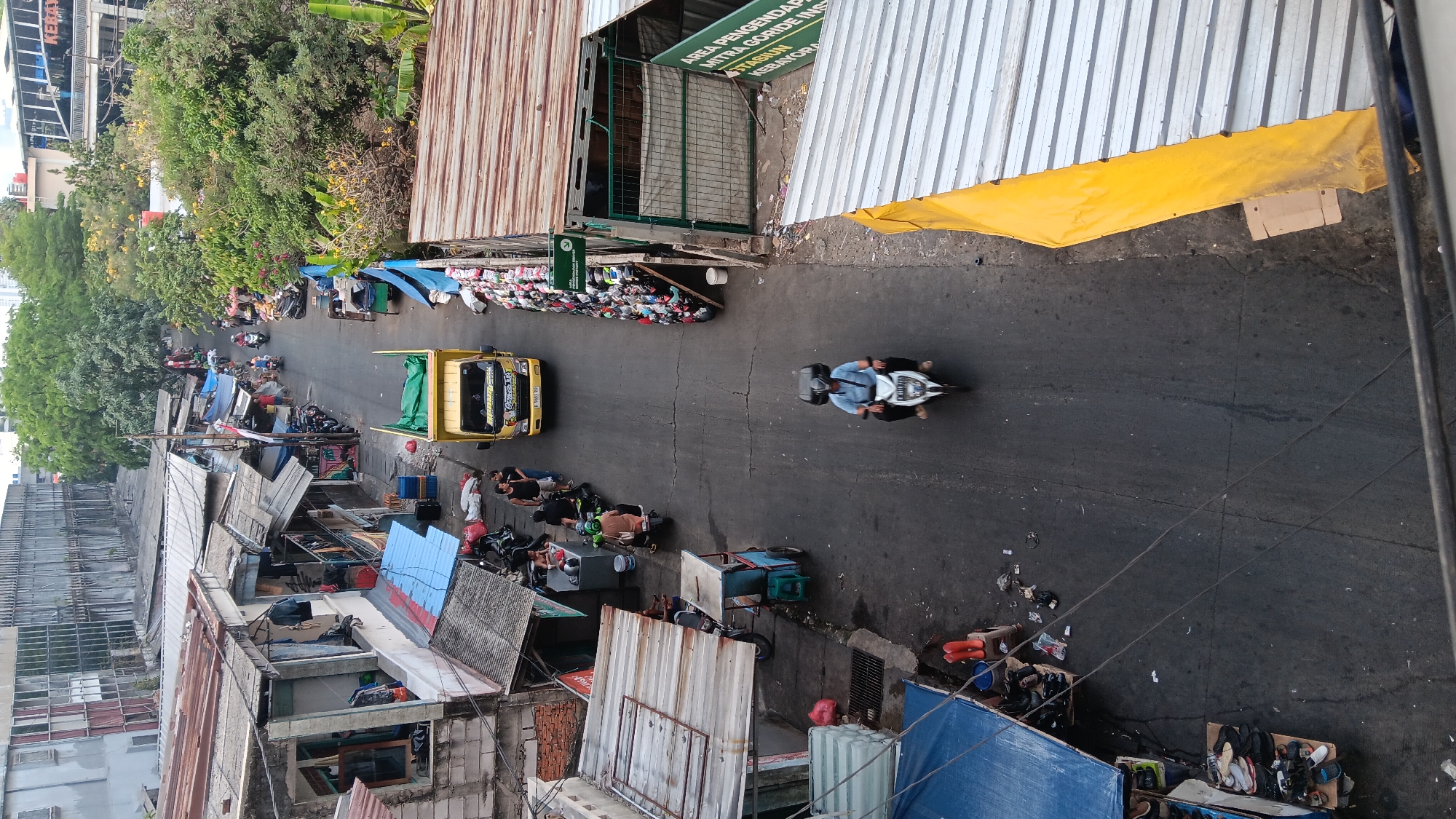 The image shows an aerial view of a narrow street in Kebayoran, South Jakarta, Indonesia, lined with informal shops and stalls. A yellow truck is parked on the right side of the street, and a person on a white motorcycle is riding away from the camera. The shops have corrugated metal roofs and are packed closely together, with various goods displayed outside. There are several other motorcycles parked along the street and a few people walking or standing around. The street is paved and appears to be in a somewhat run-down condition. Trees and other greenery are visible in the background, suggesting an urban environment.