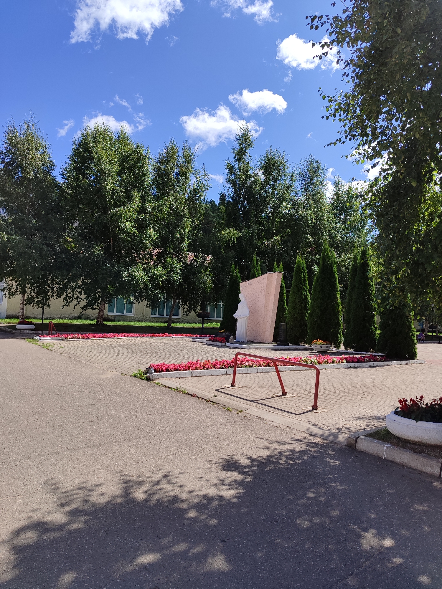 The image depicts an outdoor scene featuring a monument in a public space. In the foreground, there is an asphalt path with shadows of trees cast upon it. A red bicycle rack stands on a paved area. This paved area is bordered by flower beds filled with vibrant pink flowers. Behind the flower beds, a monument is visible, consisting of a tall, light-colored triangular slab and a white statue of a person. Several tall, dark green coniferous trees are positioned behind the monument. Further in the background, a light-colored building with numerous windows is visible, surrounded by lush green trees. The sky is blue with scattered white clouds.
