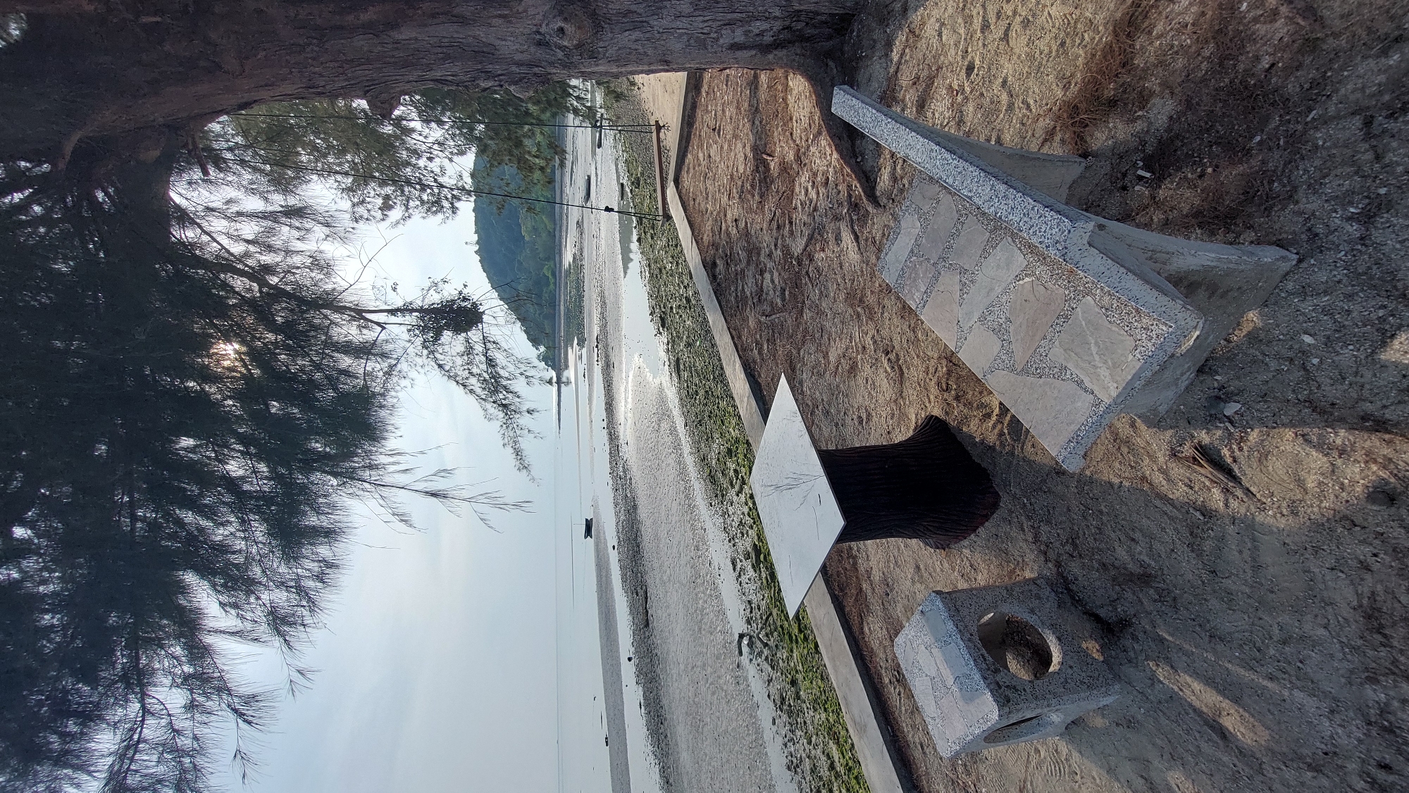 An outdoor scene in Baan Thong Po, Thailand, shows a beach at low tide. A partially visible tree stands on the left. A seating area is to the right, consisting of a stone bench, a square table with a dark textured base, and a square stone block with a circular cutout. The beach stretches toward a distant, tree-covered landmass. The sky is light, suggesting early morning or late afternoon. The tide is out, revealing sand and patches of algae or seaweed along the shoreline. Boats are visible in the distance.