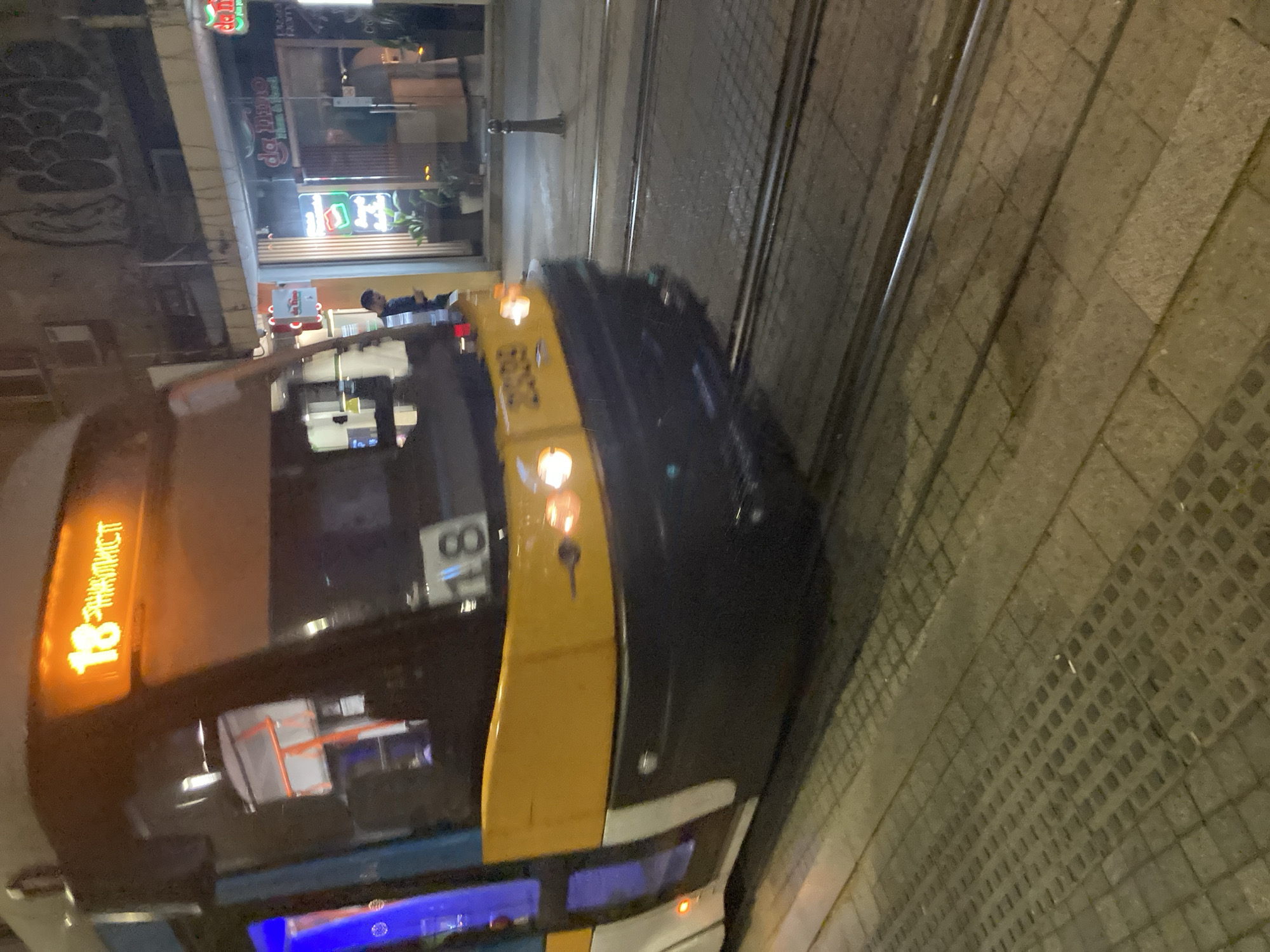 A nighttime, tilted, close-up shot depicts a Sofia public transport vehicle—a modern tram—traversing a tram track alongside a cobblestone pavement. The tram, designated "18" and labeled "STANTSIONNY KVA..." (likely referring to "Stantsionny kvartal" - Station Quarter), is partially visible, focusing on the front section. The vehicle features a color scheme of predominantly dark gray, white, yellow and blue. Reflections are visible on the tram's windows.
In the background, out-of-focus buildings are seen, including a storefront with "Da Lima" signage, suggesting a food establishment in Sofia, Bulgaria. A person can be discerned near the storefront. The tram tracks run adjacent to the pavement. The illumination is artificial, highlighting the tram and street, but leaving the surrounding areas in relative darkness.