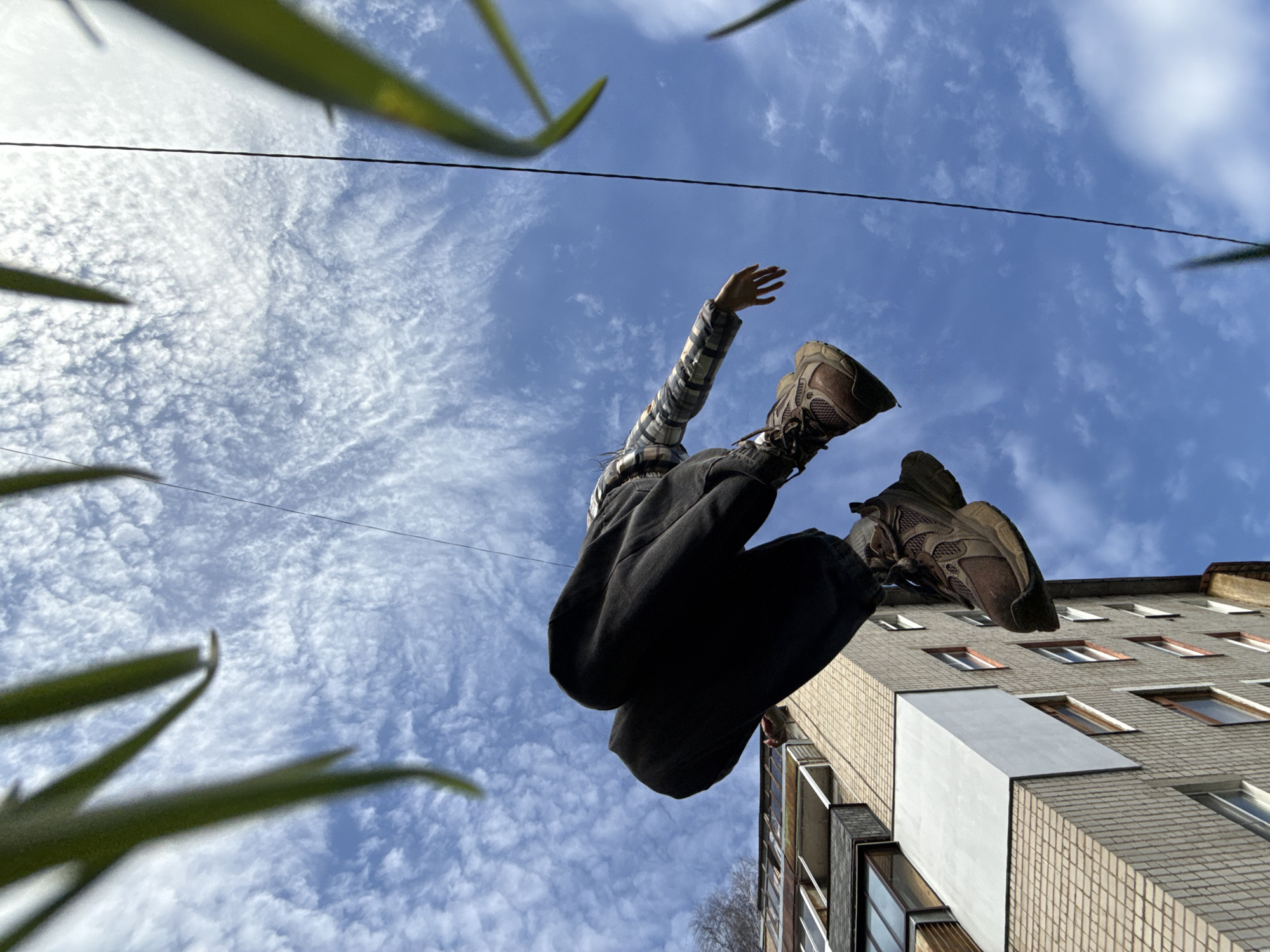 A low-angle shot captures a person suspended mid-air against a bright blue sky filled with scattered, patchy white clouds. The subject, viewed from below, is wearing a light-colored plaid shirt, dark pants, and sturdy, light brown hiking-style shoes. Their right arm is extended upwards and slightly bent, while the legs are drawn up, suggesting an energetic jump or a dynamic pose.
A thin black wire or power line stretches horizontally across the upper middle of the frame. In the extreme foreground, blurred green plant leaves frame the top and bottom left of the image, adding depth and a natural element. To the right, a multi-story apartment building with a light brown brick facade and numerous rectangular windows and balconies ascends into the frame. The building's architecture suggests an urban or suburban setting, consistent with its location in Rusanovo, Russia. The sunlight is bright, indicating daytime and clear weather. There is no visible text in the image.