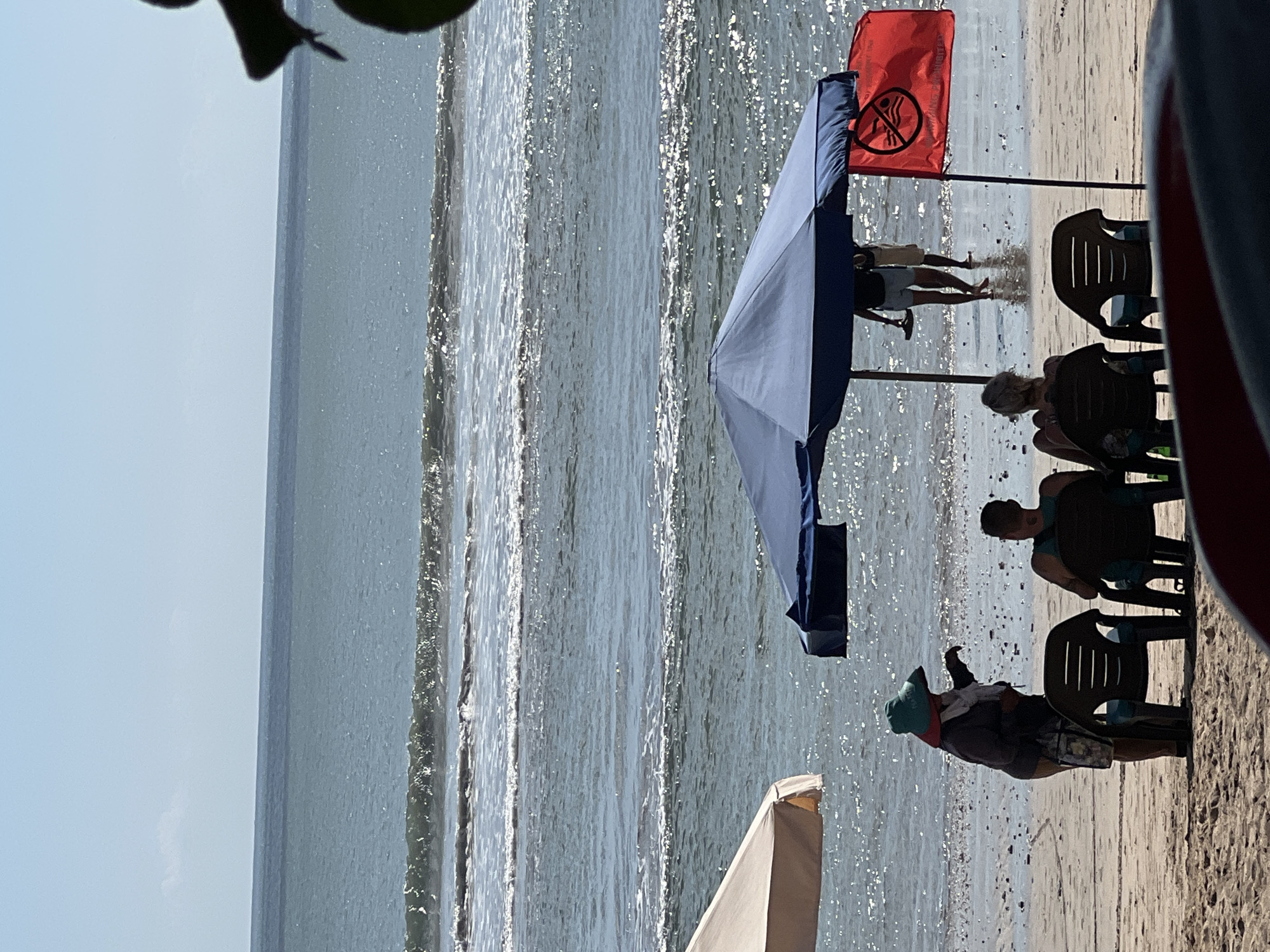 The image shows a beach scene in Kuta, Indonesia, on a bright, sunny day. The ocean is visible in the background with gentle waves. In the foreground, there are several beach chairs and umbrellas. A person wearing a wide-brimmed hat and a scarf stands near the water, appearing to interact with someone out of frame. Two people are seated in beach chairs under a large blue umbrella. Further back, two individuals are walking in the shallow water near the shore. A red flag with a "no swimming" symbol is planted in the sand. The beach appears to be sparsely populated, with the focus on the natural elements and a few beachgoers enjoying the day.