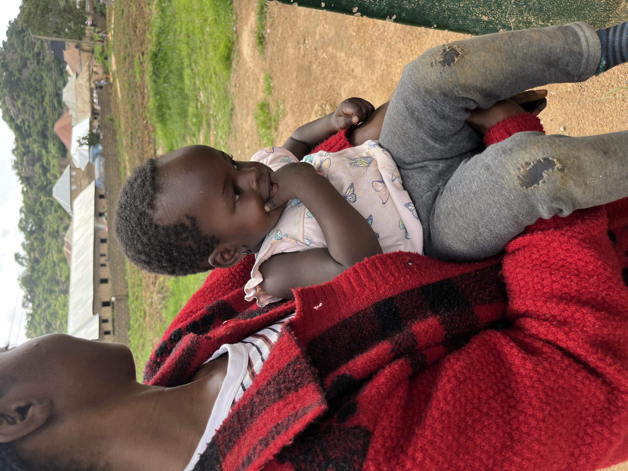 The image shows a close-up of a baby being held by an adult. The baby, facing left, has dark skin and hair, and is wearing a light-colored shirt with a butterfly pattern. The baby's right hand is near their mouth, and they appear to be looking to the left. The adult, seen from the shoulder down, is wearing a red and black plaid sweater over a white striped shirt. The adult's arm is around the baby, and their legs, clad in gray pants with visible patches, are also in the frame. The background is blurred but suggests an outdoor setting with greenery and some buildings, consistent with a rural or semi-rural environment in Bukuru, Nigeria. The lighting appears to be natural, indicating daytime. The overall mood is one of intimacy and care between the adult and the baby.