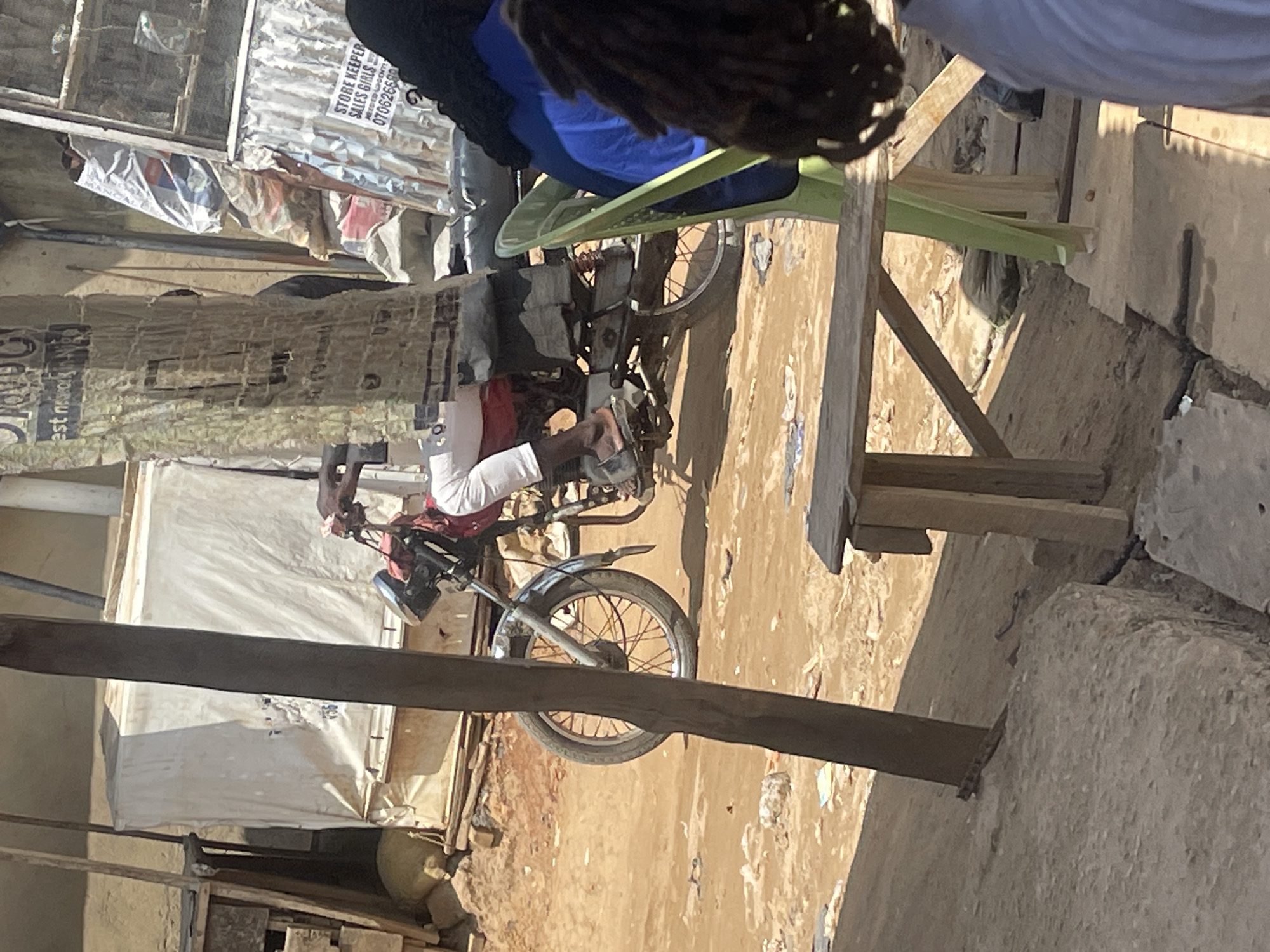 The image shows a person sitting on a motorcycle. The person is wearing white pants and a red shirt. The motorcycle is red and black. The scene appears to be outdoors, with a dusty ground and some buildings in the background. There is a large piece of corrugated metal on the left side of the image, and some wooden structures. A green plastic chair is visible behind the motorcycle. The lighting suggests it is daytime, and the weather appears to be sunny. A sign is visible on the corrugated metal, with text that reads "STORE KEEPER SALES GIRLS NEEDED URGENTLY". Below this, a phone number is partially visible. The location is identified as Abuja, Nigeria. The image is oriented vertically, with the ground appearing on the right side of the frame.