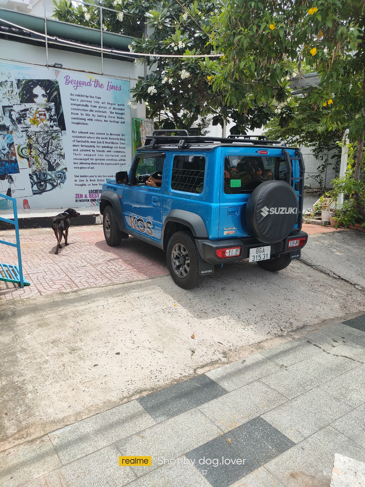 The image shows a blue Suzuki Jimny parked on a paved area. A dark-colored dog is standing on the paved area, facing away from the camera and towards the left. Behind the car and to the left is a large mural or artwork with various images and text, including the words "Beyond the lines" and "Art exhibit by Van Fran". To the right of the car, there are trees with green leaves and some yellow flowers. The ground is a mix of concrete and patterned paving stones. In the background, there is a building and some fencing. The scene appears to be outdoors in a public area, possibly a street or an outdoor dining area. The text on the mural mentions "Zen & Rest Bistro 81 NGUYEN DINH CHI" which is a location in Phu Thuy Ward, Vietnam.