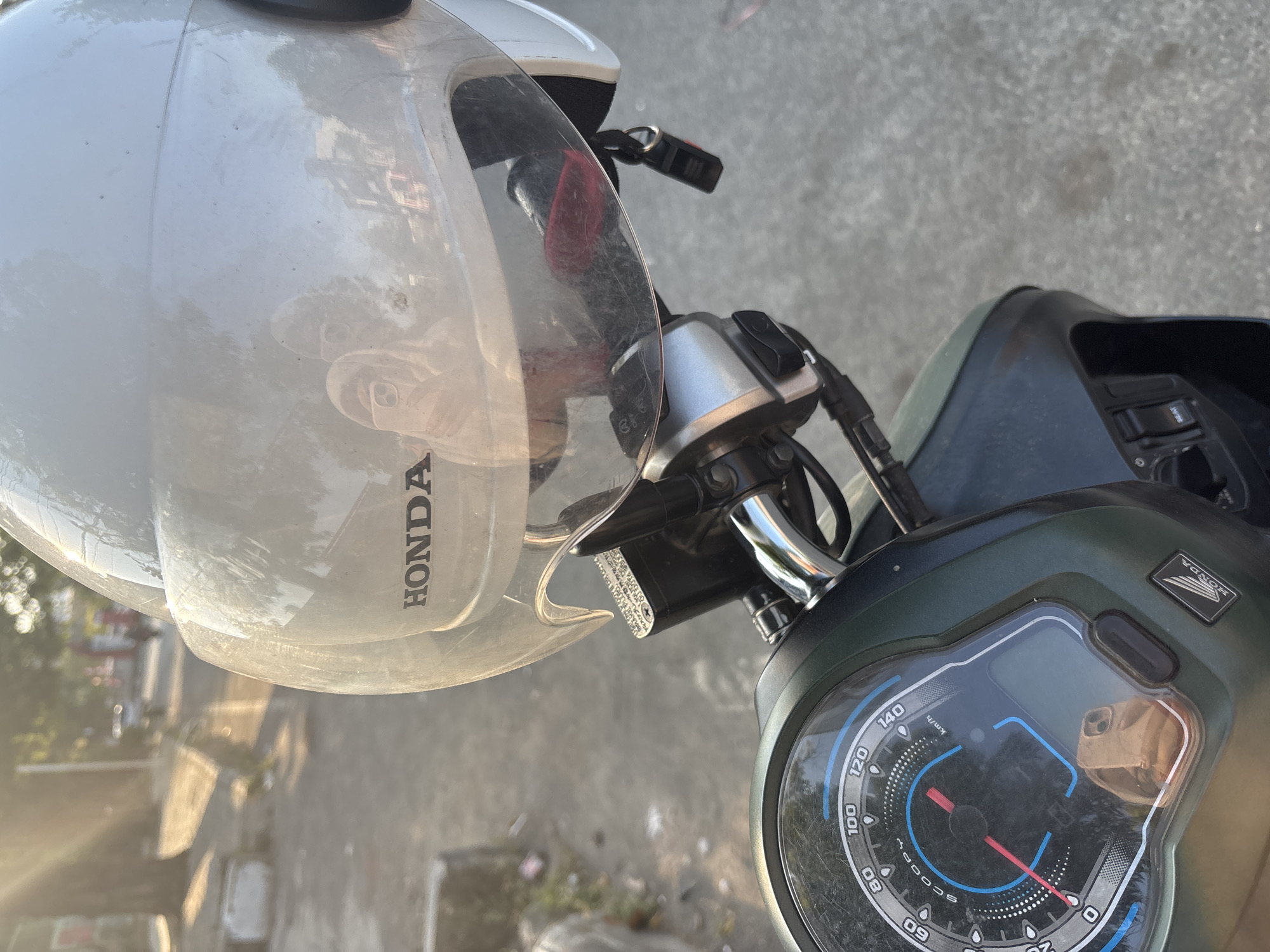The image captures a close-up view of a white Honda motorcycle helmet, positioned over the handlebars and instrument panel. The helmet's transparent visor reflects the surroundings, showing a blurry image of trees, buildings, and possibly other vehicles. The "HONDA" logo is visible on the helmet. The motorcycle's instrument cluster, featuring a speedometer that reads "SCOOPY," is also in focus. The background suggests an outdoor setting, likely a street or parking area, with a paved surface and some vegetation. Sunlight casts a warm glow on the scene, creating lens flare effects. The scene appears to be in Cirebon, Indonesia, based on the motorcycle model and general context of scooter use in the region.