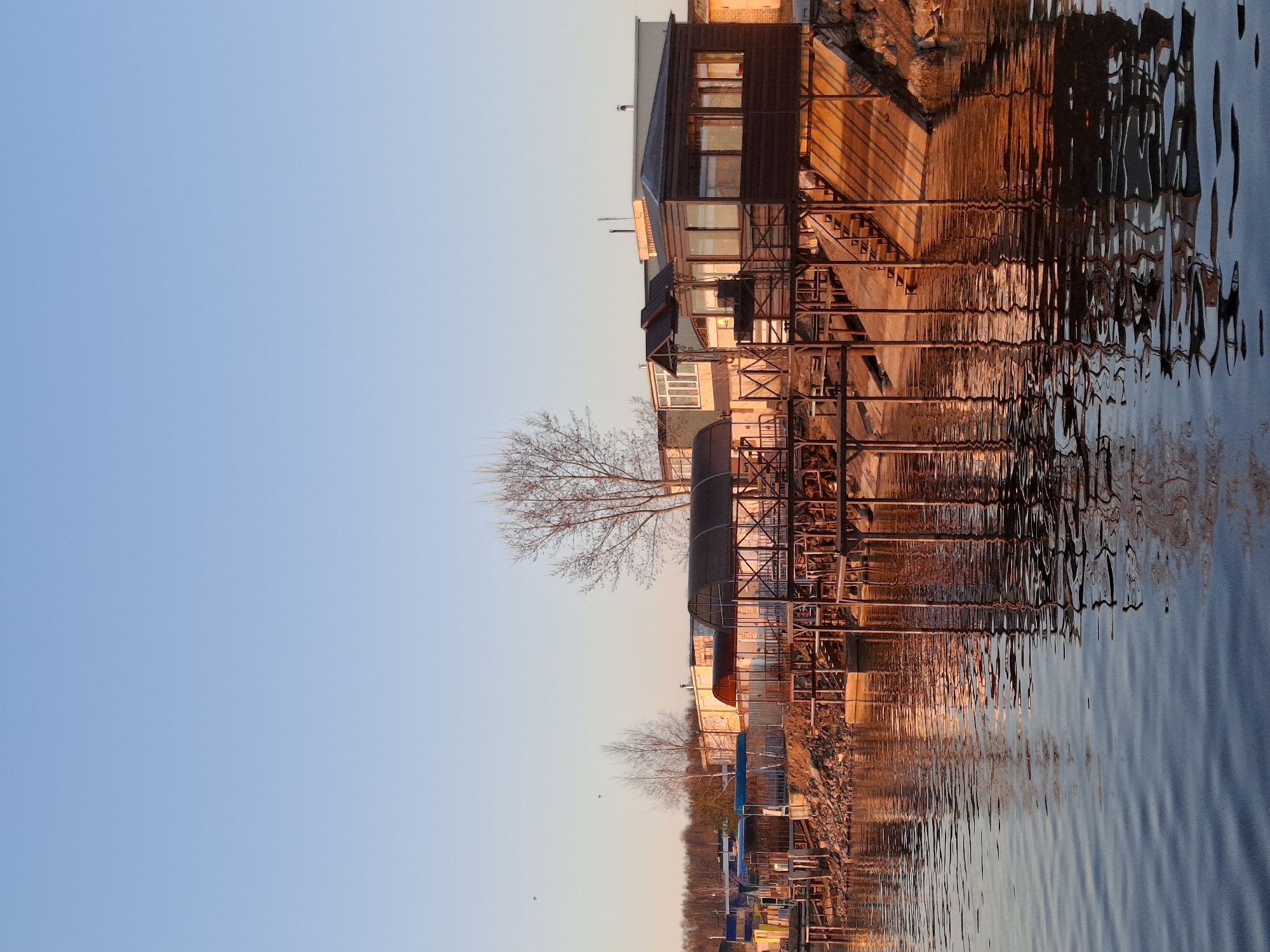 The image depicts a waterfront scene in Chaikovsky, Russia, likely at sunset or sunrise based on the warm golden light. A multi-story building, possibly a house or recreational facility, sits partially over the water supported by a steel framework. The building has a dark facade with numerous windows. The water reflects the building and framework in distorted patterns. Bare trees are visible next to the building and along the shoreline. In the foreground, a row of boats or docks extends into the water. The sky above is clear and blue.