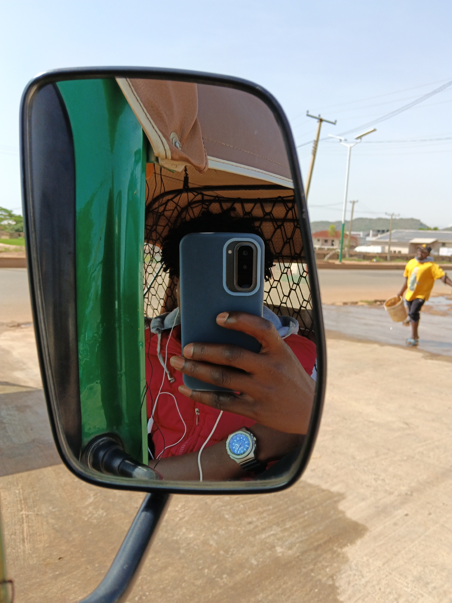 The image is a first-person perspective selfie taken in the reflection of a vehicle's side mirror. The mirror shows the photographer holding a dark blue smartphone, capturing their own reflection. The photographer is wearing a red jacket and white earphones. A silver watch with a blue face is visible on their left wrist. In the background, reflected in the mirror, is a person in a yellow shirt and black shorts walking on a dirt road, carrying a bucket. The environment appears to be outdoors, possibly a street or roadside, with utility poles and buildings visible in the distance. The overall lighting suggests it is daytime.