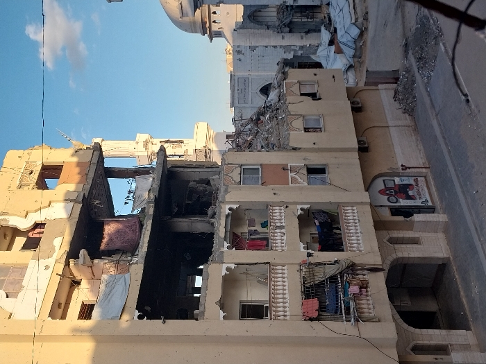 The image depicts a multi-story residential building in Khan Yunis, Palestinian Territory, exhibiting extensive structural damage. The upper three floors are largely destroyed, with external walls blown out, exposing internal rooms, rebar, and rubble. The facade shows visible shrapnel or bullet impacts. On the lower, relatively less damaged floors, multiple balconies feature clotheslines with laundry, including various garments and a prominent red patterned rug hanging from a mid-level damaged balcony. An exposed interior of a room on a lower floor contains additional hanging laundry. At ground level, an archway leads to an interior space, and a storefront sign for "HASHEM GAMES" with "Squid Game" graphics (red figure, circle, triangle, square) is visible next to an entrance. Air conditioning units are mounted on the building's exterior.
To the right of this building, a large mosque is visible, also appearing damaged with rubble on its roof and a visible minaret on its left side. Further right, another damaged building is partially visible. The immediate foreground features a paved street with scattered debris and piles of rubble at the base of the buildings. White tarpaulins cover additional debris near the mosque. The sky is clear blue with scattered white clouds, illuminated by daylight. No individuals are visible in the scene.