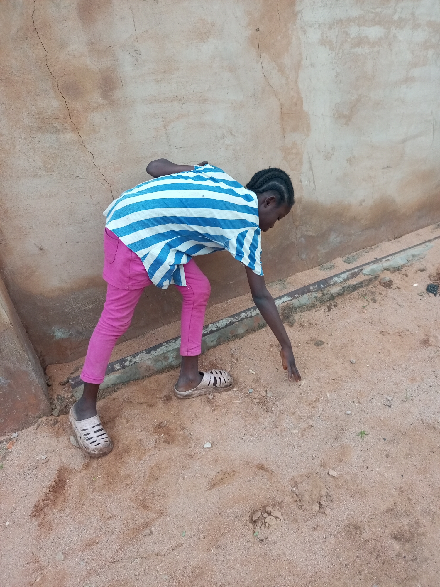A young girl with braided hair, wearing a blue and white striped shirt and pink pants, bends down to touch the sandy ground in front of a textured, tan-colored wall. She is wearing light-colored sandals. The ground is composed of sand and small pebbles. The environment appears to be outdoors, possibly in an urban setting within Tempure, Nigeria, as indicated by the wall and the sandy ground.